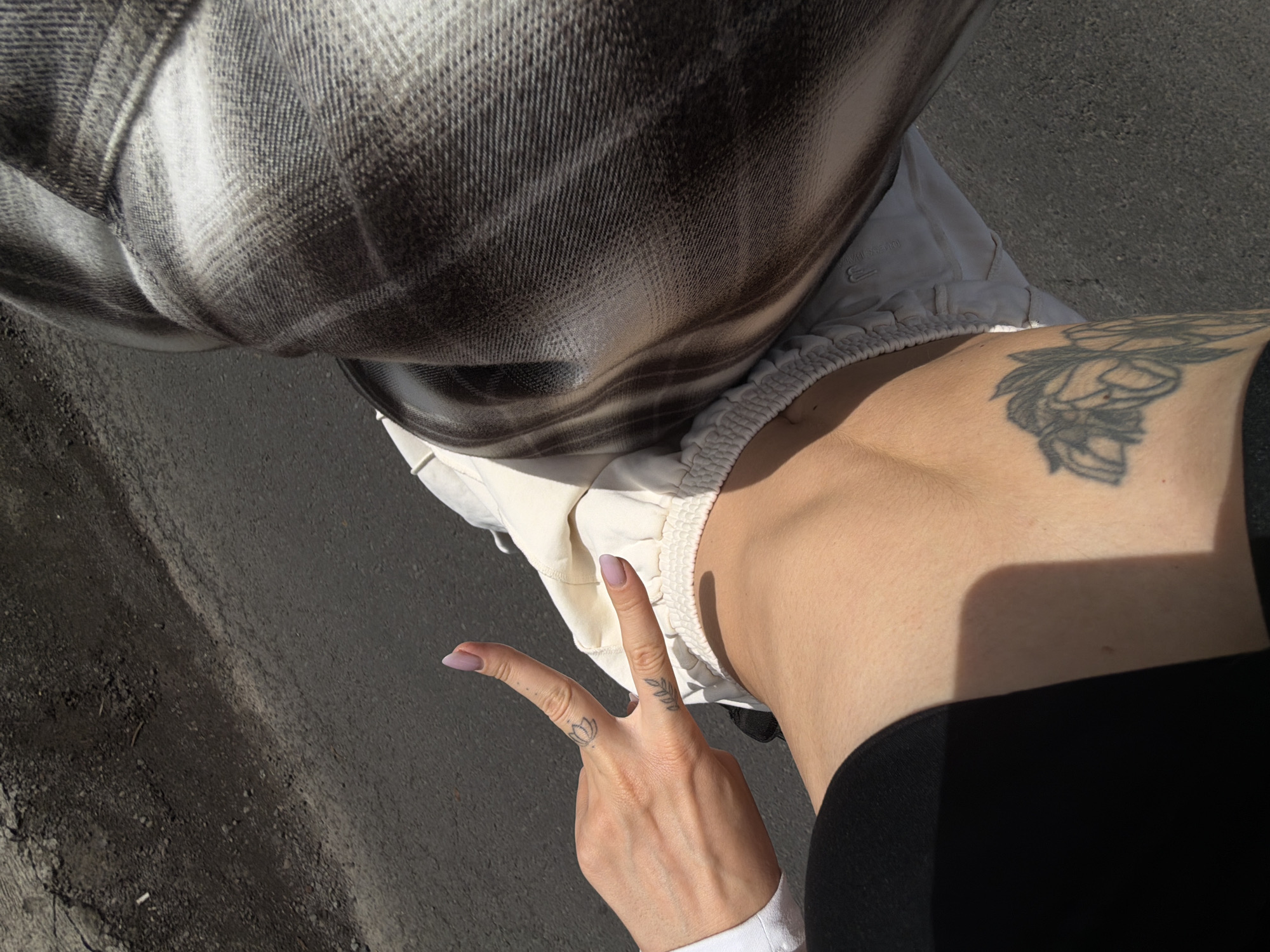 A close-up, high-angle shot shows a person's hand and leg from the perspective of someone looking down at their own body. The person's left hand is raised, fingers spread in a peace sign, with two small tattoos visible on the knuckles and the side of the index finger. The fingers are painted with a light purple nail polish. The person is wearing light-colored sweatpants with a drawstring waist and a black garment on their lower left side. A large tattoo of a floral design is visible on the person's thigh. The background consists of asphalt, suggesting an outdoor urban or suburban environment. The lighting indicates it is daytime with a shadow cast on the person's leg. The location is identified as Mengwi, Indonesia.