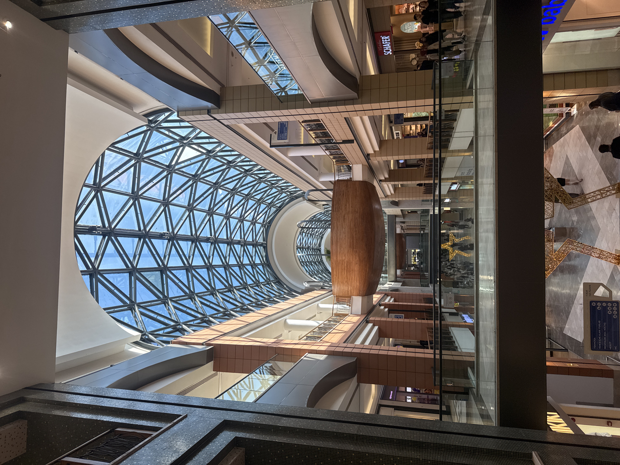 This image captures the vibrant interior of a multi-story shopping mall, likely located in Bayrampaşa, Turkey. The most striking architectural feature is a massive, circular, geodesic glass skylight that forms a domed ceiling, allowing abundant natural light to illuminate the space. Below the skylight, a large, ornate wooden cylindrical structure serves as a central design element.
The mall features multiple levels with balconies and walkways, all overlooking a central atrium. Numerous people are visible across different floors, walking and congregating, indicating a lively shopping environment during daytime hours due to the bright, sunlit interior.
Notable details include a large, golden, decorative structure resembling the Eiffel Tower on a lower level, and a glowing golden star decoration on an upper level. The mall's design incorporates modern aesthetics with glass railings, tiled surfaces, and warm wooden accents.
Visible text includes store names like "SCHAFER" on a red sign on an upper floor. A blue directional sign on a lower level provides information, listing "1st FLOOR" (with "LKAT" possibly being a Turkish equivalent for floor), "TEKNOSA," "YEME & İÇME ALANI FUNLAB," "DECATHLON," "IKEA," "KOÇTAŞ," "MESCİT" (prayer room), and "WC" (restroom), each accompanied by icons and "000," likely indicating floor numbers or service areas. Other partial text like "IKIN" and the number "383" are also present.