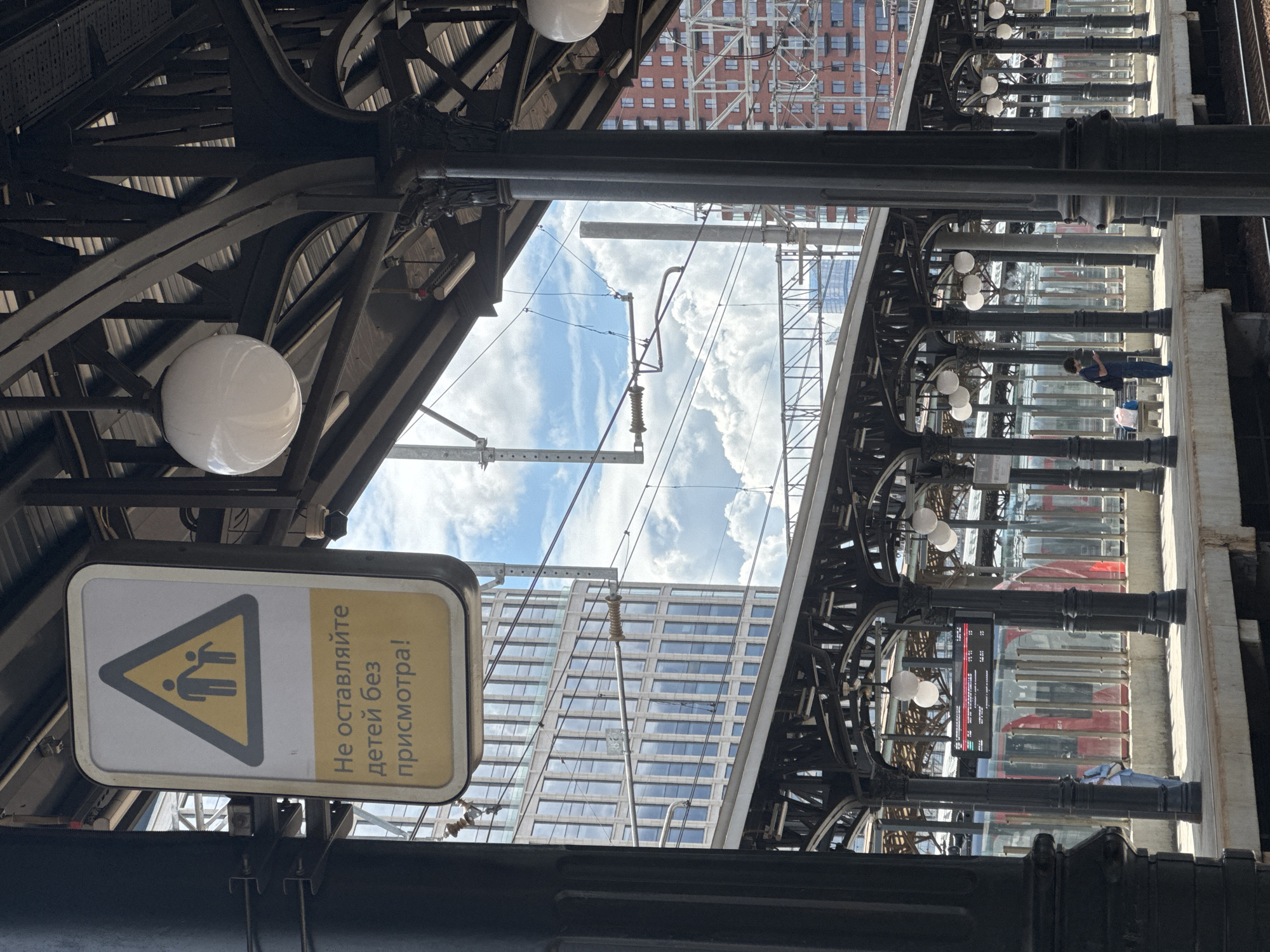 The image depicts a view from a train platform at a station, likely in Moscow, Russia, indicated by the Russian text on the warning sign. A black metal pillar is prominent in the foreground, with a sign attached to it that reads "Do not leave children unattended!" in Russian, accompanied by a yellow triangular warning symbol featuring silhouettes of an adult and a child. Above, a white spherical light fixture is visible, and the structure of the station roof is made of dark metal beams. Beyond the platform, a train with red and white elements is partially visible. In the background, a modern glass-facade skyscraper and a red brick building are seen against a sky filled with cumulus clouds. On the platform, a person wearing a blue shirt and dark pants is standing near a bench, looking at their phone, with a bag beside them. Another person, wearing a light blue top and dark pants, is standing further down the platform. Electrical wires and supporting structures for the overhead catenary system are visible crisscrossing the sky.