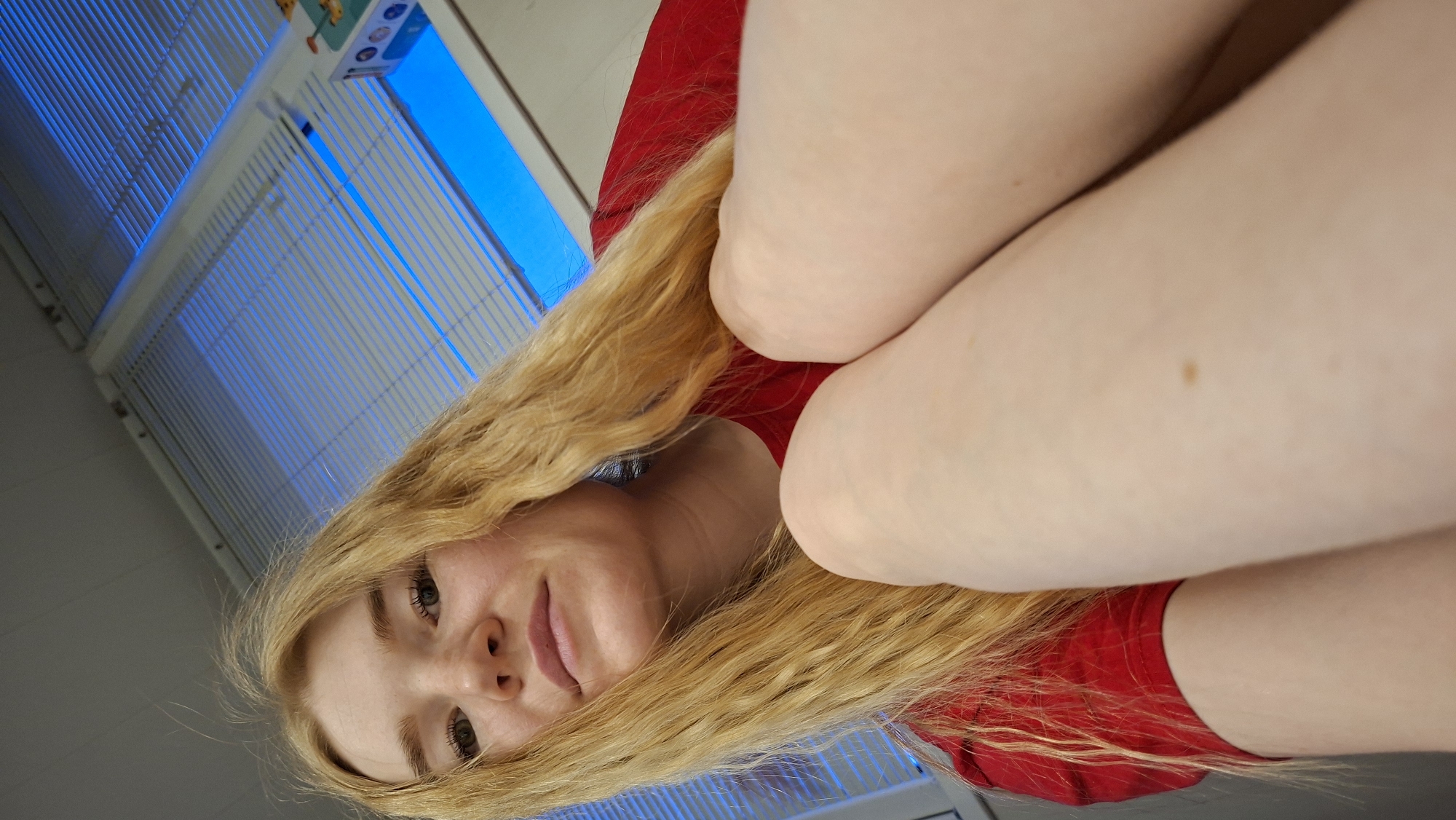 A close-up, low-angle portrait features a young woman with long, wavy blonde hair, looking directly at the camera with a soft, neutral expression. Her fair-skinned face occupies the lower left portion of the frame, with her light-colored eyes and slightly parted lips visible. She is wearing a red top. Her bent arms or legs (likely upper legs/thighs) are prominently positioned in the foreground, creating a partial obstruction and appearing fair-skinned, with a distinct brown mole visible on one limb.
The background consists of an indoor setting, likely a room in Chaikovsky, Russia, with a white ceiling. Fluorescent light fixtures with grilles run across the ceiling. One fixture emits a strong blue light, casting a distinct cool tone on that section of the ceiling. A small, rectangular object with indistinct colorful shapes and possible, but illegible, text is visible near the blue light fixture. The overall lighting suggests an artificial indoor environment. The woman's pose and direct gaze suggest a self-portrait or a very close-up photo taken from a low perspective, possibly while she is reclining or sitting.