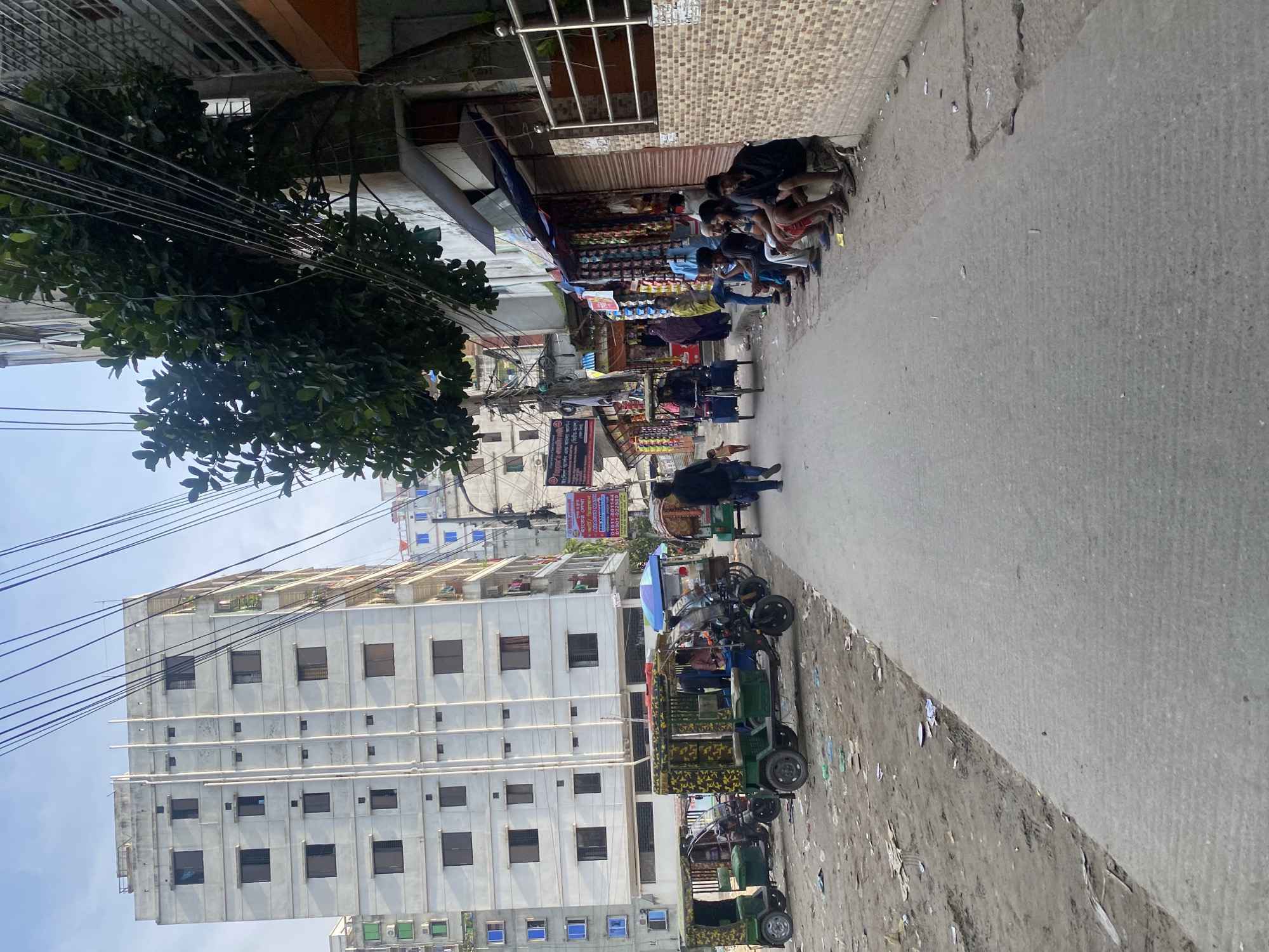 The image depicts a street scene in Dhaka, Bangladesh. In the foreground, a paved road runs diagonally from the bottom right towards the upper left. To the left of the road is a dirt lot with several auto-rickshaws parked, some covered with camouflage-patterned tarps. Further down the street on the left, a tall, multi-story concrete building with numerous windows dominates the background. On the right side of the road, a row of shops with awnings is visible, displaying various goods. Several men are seated on a low wall in front of the shops, and a few people are walking along the street. Trees and utility wires are prominent features of the environment. The scene appears to be a busy urban area, likely a marketplace or transportation hub.