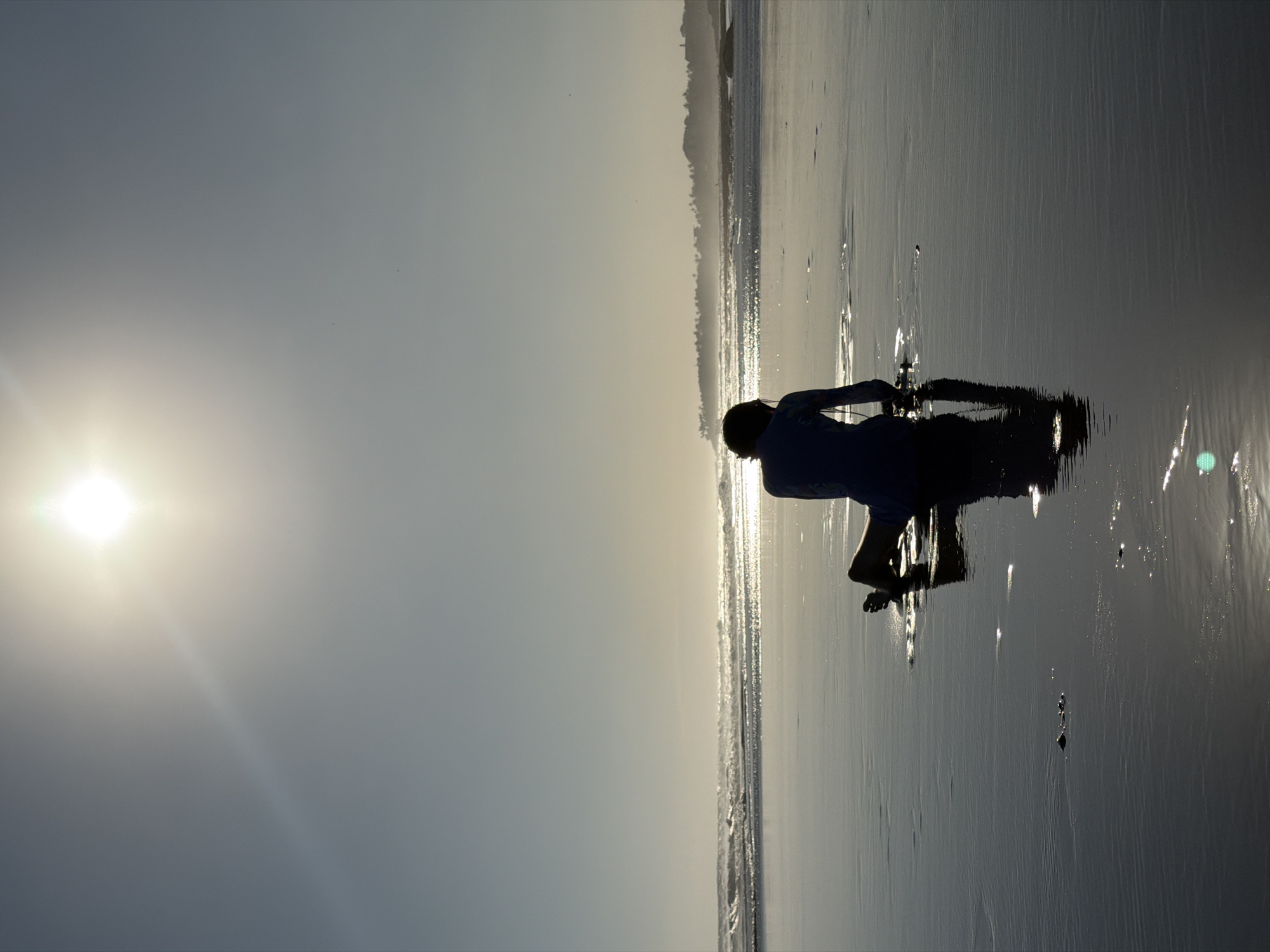 The image captures a silhouetted figure kneeling on a wet sandy beach in North Kuta, Indonesia. The sun, positioned high in the sky, creates a strong glare and casts a reflection on the waterlogged sand. The figure is facing away from the camera, towards the ocean, where gentle waves are breaking. In the distance, a hazy line of trees and structures can be seen against the horizon, typical of a coastal landscape in the region. The overall atmosphere is serene and backlit by the bright sun.