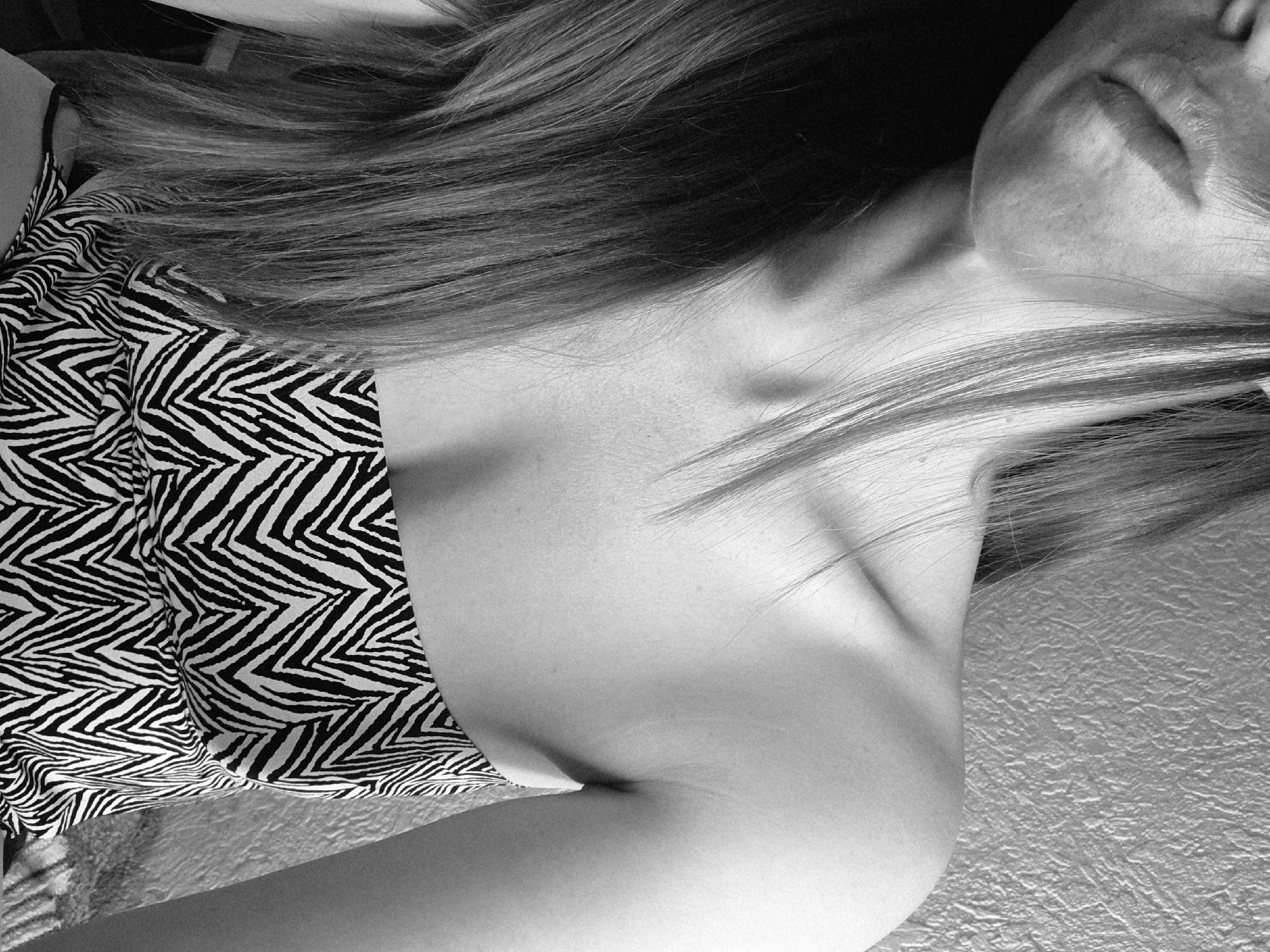 A black and white photograph shows a portion of a person's torso, neck, and face. The subject is wearing a top or dress with a black and white zebra-striped pattern. Hair is draped across the shoulder and chest area, partially obscuring the neck. The face is partially visible, showing the lips and part of the nose. The background appears to be a textured wall. The image does not provide information to ascertain its location in Chaikovsky, Russia, or information about actions, interactions, or additional subjects or objects.