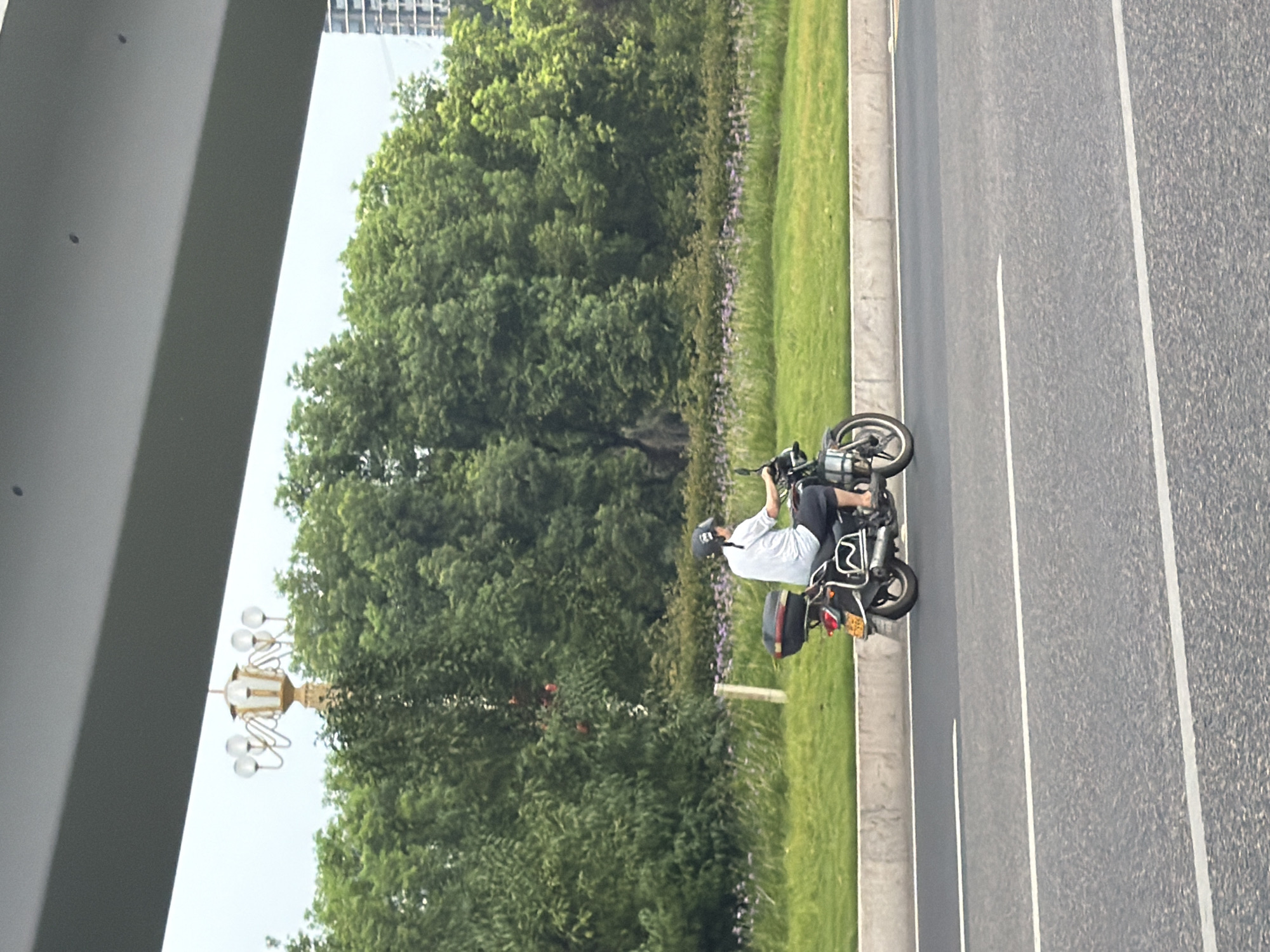 A person wearing a helmet and a light-colored shirt is riding a motorcycle on an asphalt road. The motorcycle has a black storage box attached to the back. The road has white lane markings. To the right of the road, there is a grassy median with a curb, and beyond that, a dense line of lush green trees. In the background, a modern building with a grid-like facade is visible. Above the trees, a decorative lamppost with multiple globes is perched on a golden pole. The sky is overcast. The image appears to have been taken from a moving vehicle. This scene is located in Jiangjin, China.