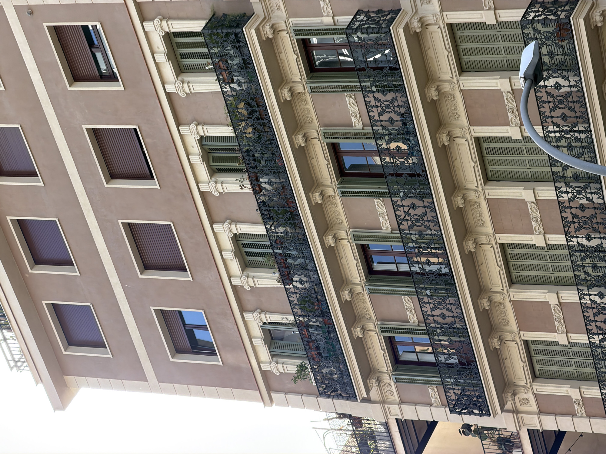 This is a low-angle, exterior, full-shot photograph of a building facade. The building has a pale pinkish-brown exterior with white trim. There are multiple windows with dark brown shutters and some with visible windows reflecting the sky and the building opposite. Each floor features a wrought-iron balcony with an intricate, decorative pattern. The balconies have some greenery and potted plants. The architectural style appears ornate, with decorative moldings and columns between the windows. A modern, gray street lamp curves into the frame from the right side of the image. The lighting suggests it is daytime, likely midday given the brightness. The angle of the photograph makes the building appear to tilt upwards, creating a slightly dramatic perspective. The scene is likely a street in a European city, given the architecture and the presence of a street lamp. No people are visible. There is no visible text in the image.