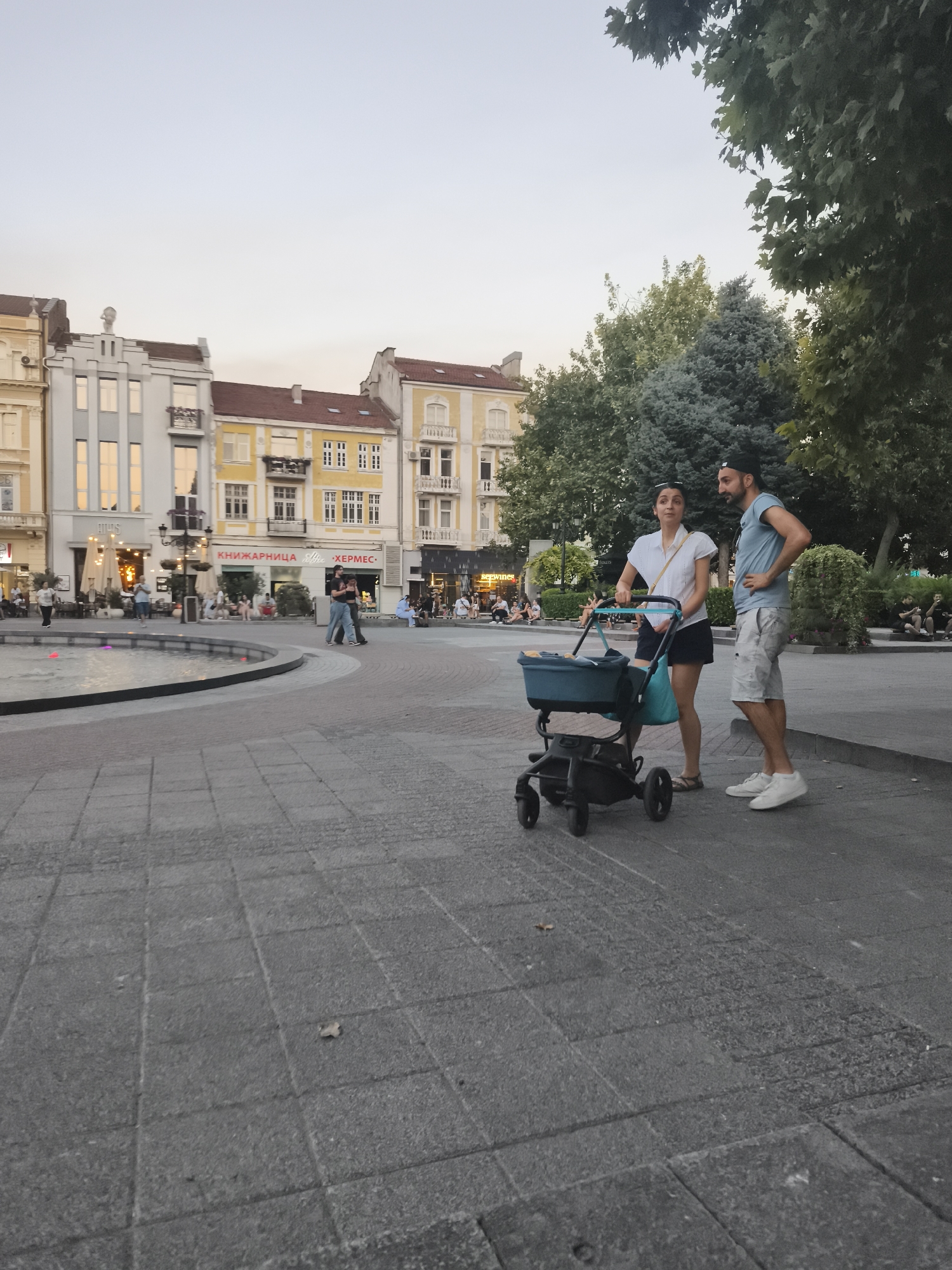 The image depicts a public square in Plovdiv, Bulgaria, likely the central pedestrian zone or a main square. In the foreground, a couple stands near a baby stroller. The woman, wearing a white shirt and dark skirt, holds the stroller's handle. The man, in a blue t-shirt and light shorts, stands beside her, looking to his left with his hands on his hips. The stroller is teal and black. The ground is paved with grey square tiles. In the background, a fountain with colored lights is partially visible on the left. Behind the fountain and square are several multi-story buildings with classical architecture, painted in light yellow and white, with red-tiled roofs. The sky is a pale, hazy blue, suggesting either early morning or late afternoon light. Trees with green foliage are present in the mid-ground and background on the right side of the image, framing the buildings and the open square. Other people are seen in the distance, seated or walking, and some outdoor seating areas are visible in front of some buildings.
