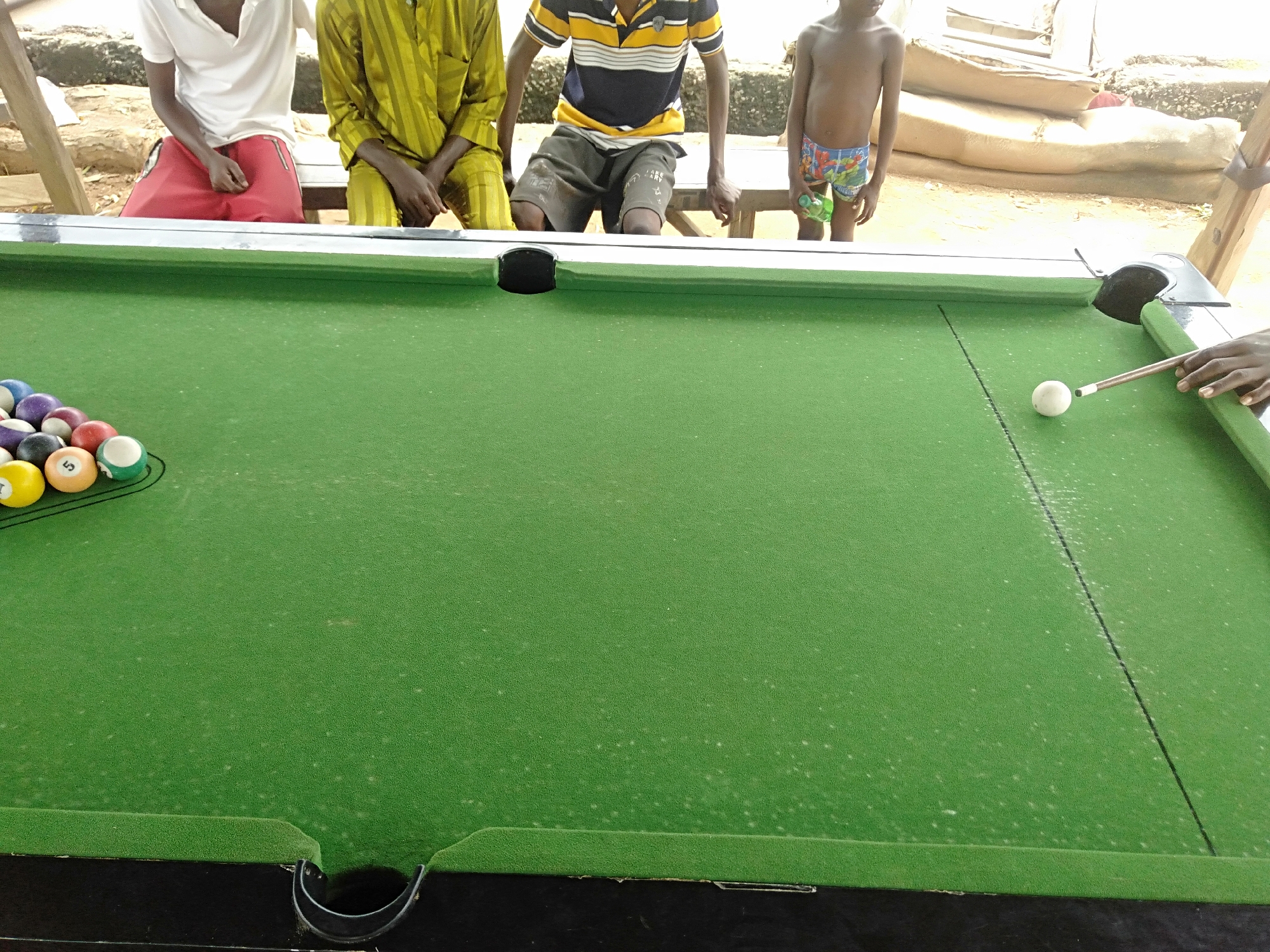 The image depicts a billiards table with a set of pool balls racked on the left and a cue ball positioned on the right, near a person holding a cue stick. The table is covered in green felt and has visible pockets at the corners and possibly along the sides. Four people are situated behind the table: one wearing a white shirt and red shorts, one in a yellow striped garment, another in a navy and yellow striped shirt and gray shorts, and a young shirtless child in patterned swim trunks holding a bottle. The location appears to be outside in an urban area, with indeterminate terrain behind the people.