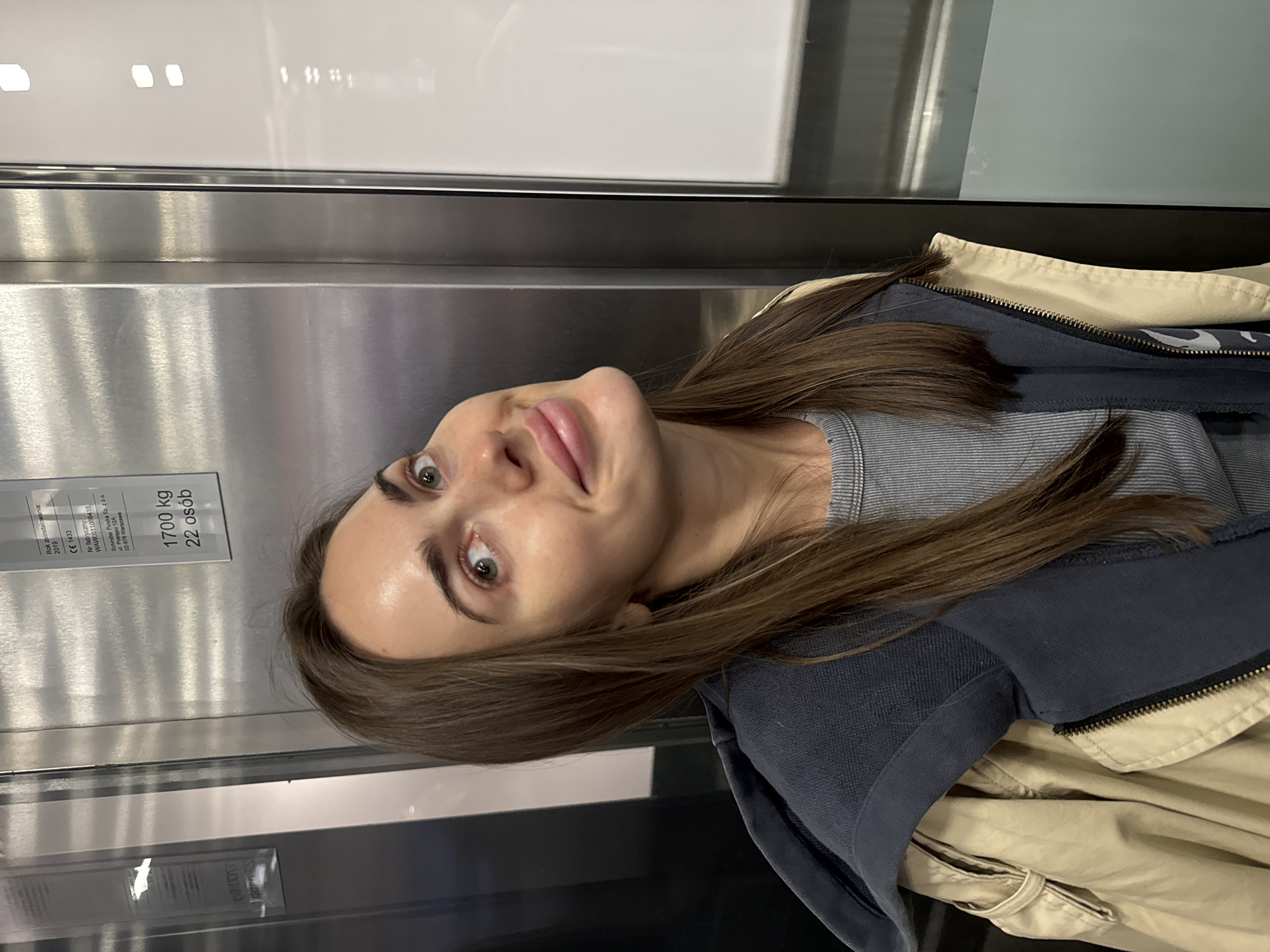 A woman with long brown hair is taking a selfie in an elevator. The elevator appears to be made of stainless steel. There is an information placard that is partially visible, with text written in Polish, and shows "Rok zainstalowania: 2019," "1700 kg, 22 osób." The woman is wearing a striped top, a dark blue zip-up jacket, and a tan trench coat.