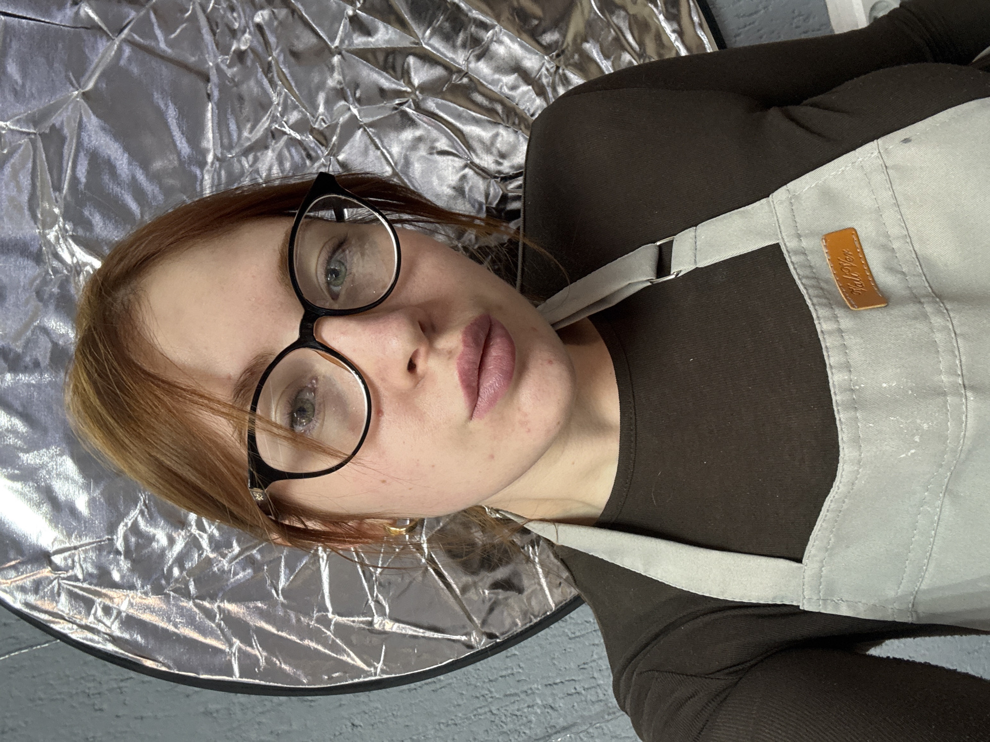 A female individual with reddish-brown hair is depicted in a close-up, facing the camera directly. She wears black-framed glasses and a dark brown long-sleeved shirt. A light grey apron, visibly smudged with white residue, is worn over the shirt, featuring a small tan rectangular patch on the upper left chest area with indistinct text. A gold hoop earring is visible in her right ear. Her lips are coated with a muted pink-purple lipstick. The background features a textured, muted blue-grey wall. To the subject's left side (viewer's right), a circular silver photography reflector with a crumpled surface is partially visible, indicating an indoor setting. The precise location within Chaikovsky, Russia, cannot be determined from the visual evidence presented.