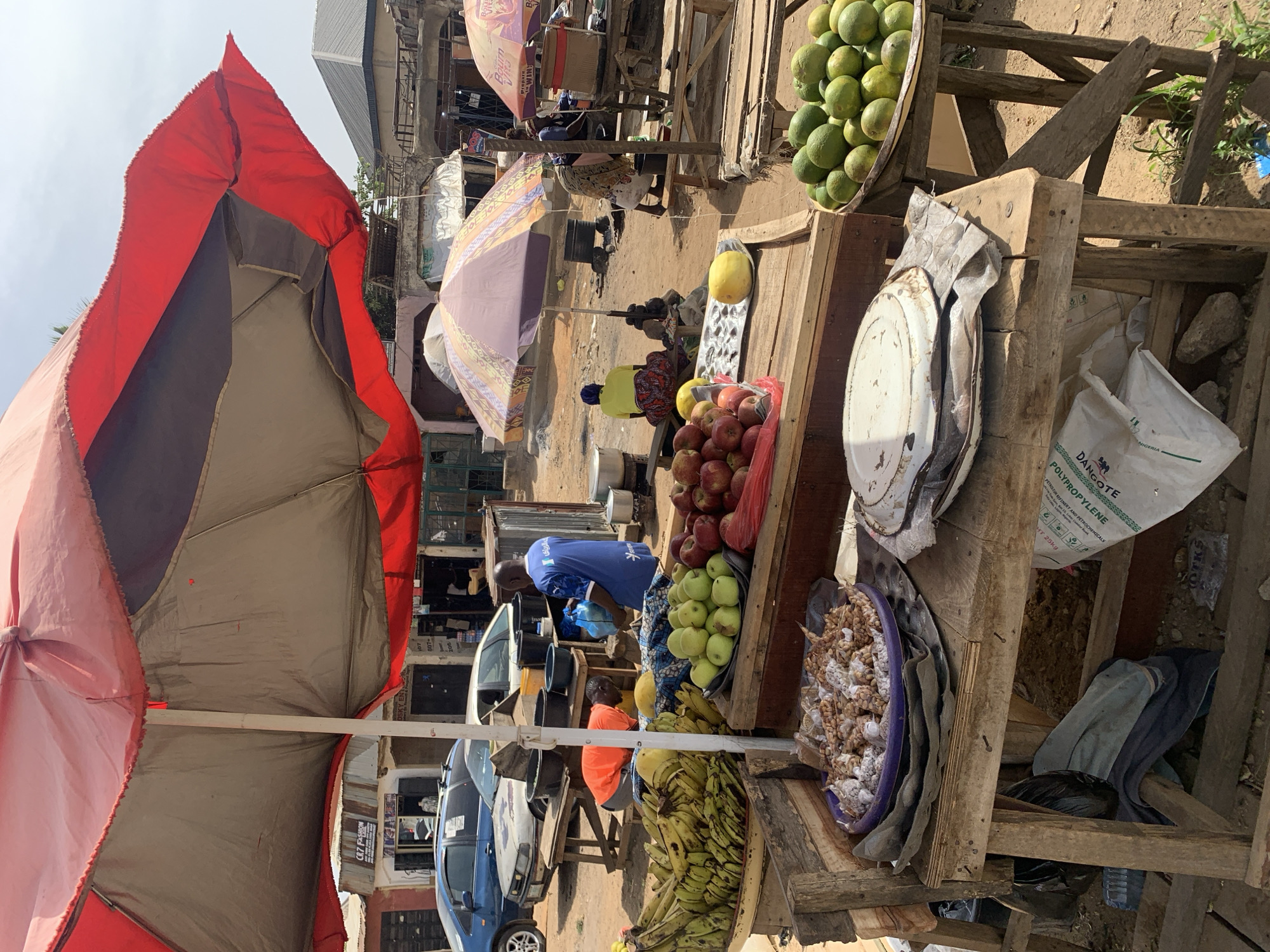 The image depicts a bustling outdoor market scene in Rafin Par, Nigeria. Wooden stalls display a variety of produce, including apples, bananas, mangoes, and peanuts in a blue bowl. Some of the items are situated on wooden racks. There is a white plastic bag with “Dangote Polypropylene” printed on it.
People are visible in the background, with at least two vendors manning their stalls. One vendor wears a blue shirt while another wears orange. The market is shaded by several umbrellas in red and purple.
Cars are parked on the side.