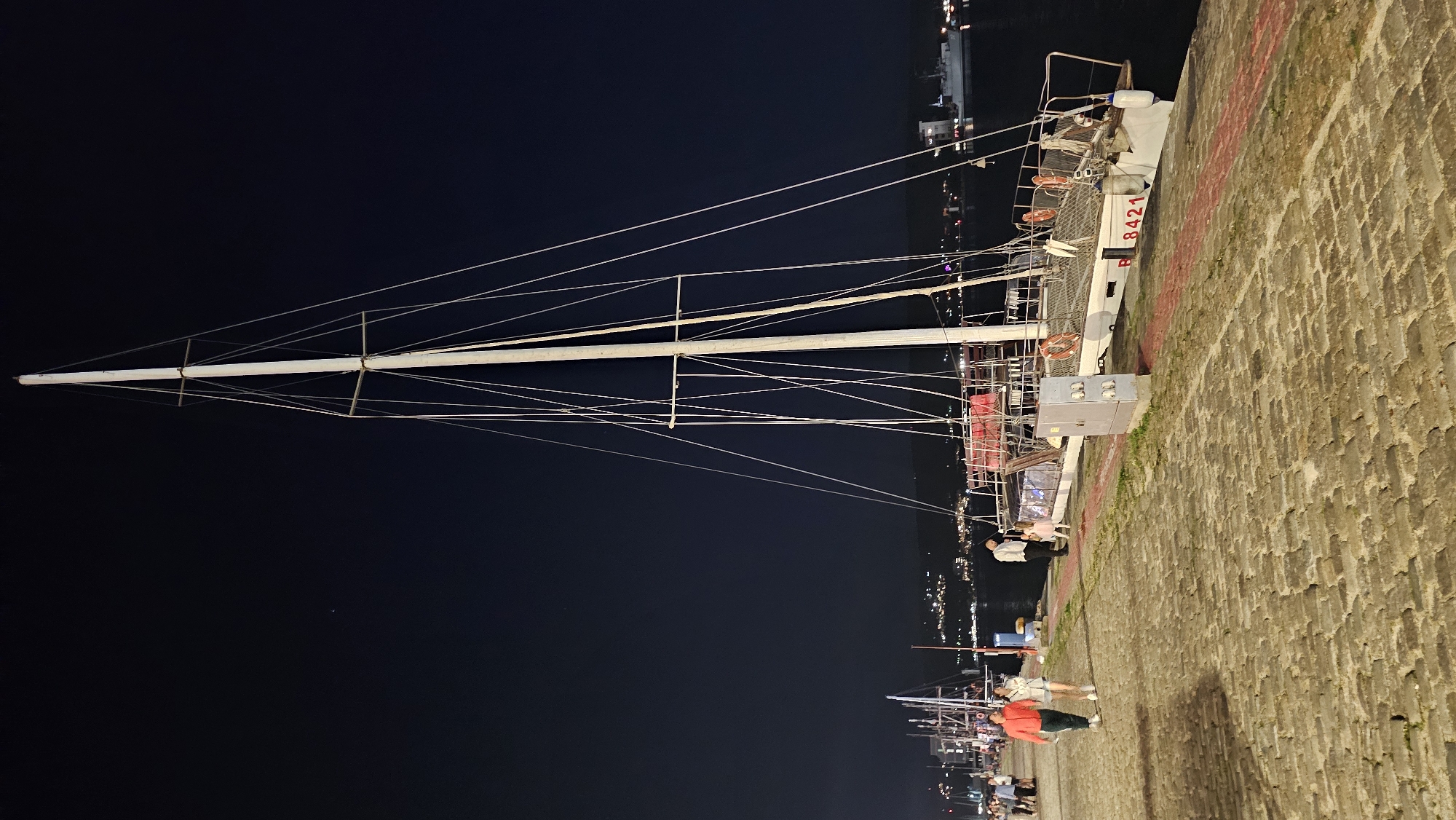 The image captures a nighttime scene at the Varna Sea Garden's pedestrian promenade, overlooking the Varna Bay. A tall sailboat with a white mast and numerous rigging lines dominates the right side of the frame. The boat, identified by the registration number "B 8421" on its hull, is docked alongside the cobblestone promenade. Several life rings are visible on the deck.
On the promenade, three people are visible. Two individuals, a man in an orange jacket and a woman in a light-colored top and shorts, walk away from the camera towards the left. Another person, a man in dark clothing, stands near the sailboat, interacting with a child in a pink outfit. Further in the background, other boats and distant city lights are discernible. The sky is dark and clear, with a faint celestial body visible. The ground is paved with rough, interlocking cobblestones, and a red-lined border runs along the edge of the promenade next to the water.