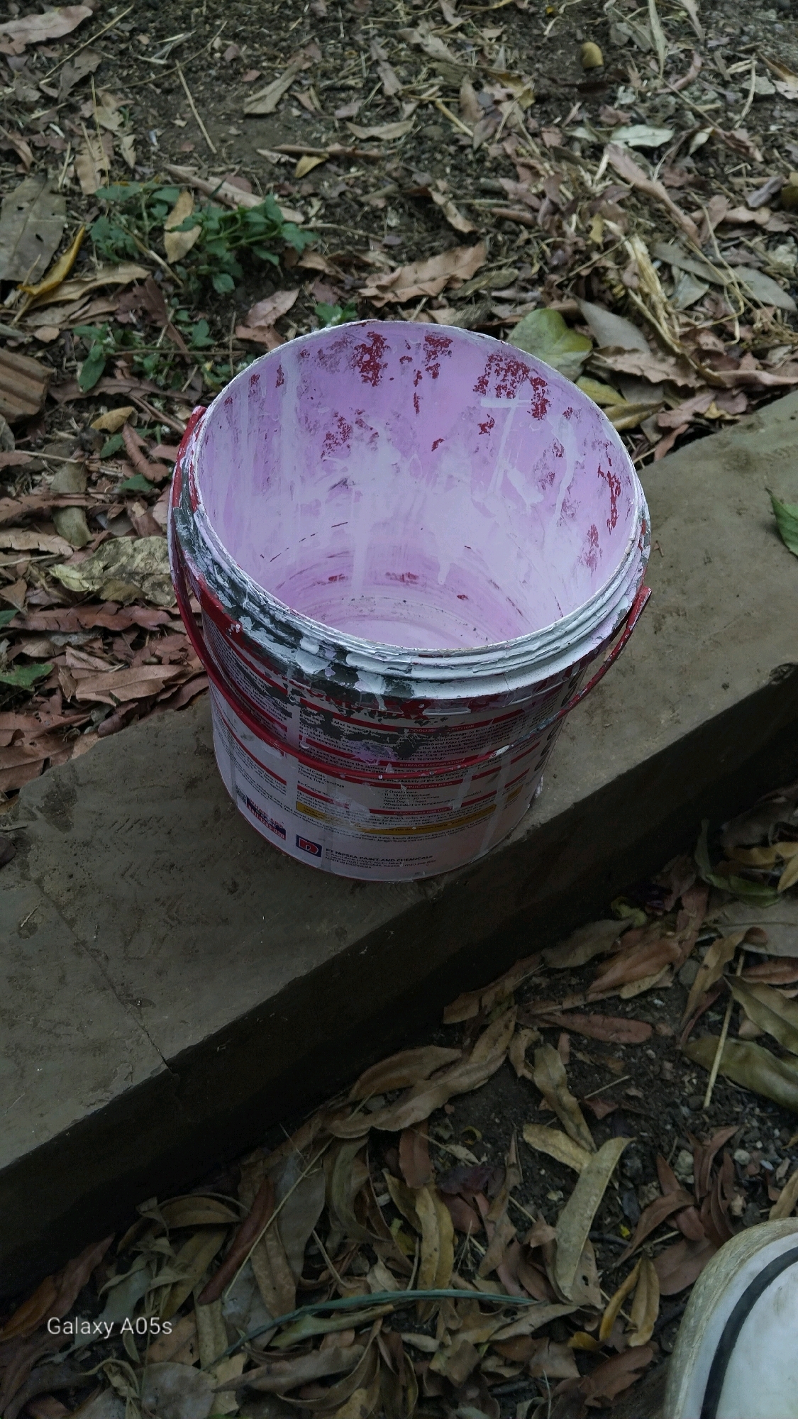 A close-up, slightly overhead shot shows a used, open paint bucket with dried pink paint splatters inside and on the rim. The bucket, which appears to be made of metal or thick plastic, has a red handle and is partially covered with a label that has red and white text. It is sitting on a rough, grey concrete surface, possibly a low wall or ledge. The background is out of focus, showing dry leaves, dirt, and some sparse green foliage, suggesting an outdoor setting. The lighting is somewhat dim, indicating it might be overcast or late in the day. In the bottom left corner, the text "Galaxy A05s" is visible, indicating the device used to capture the image. The scene is likely situated in Sumedang, Indonesia, given the information provided in the prompt.