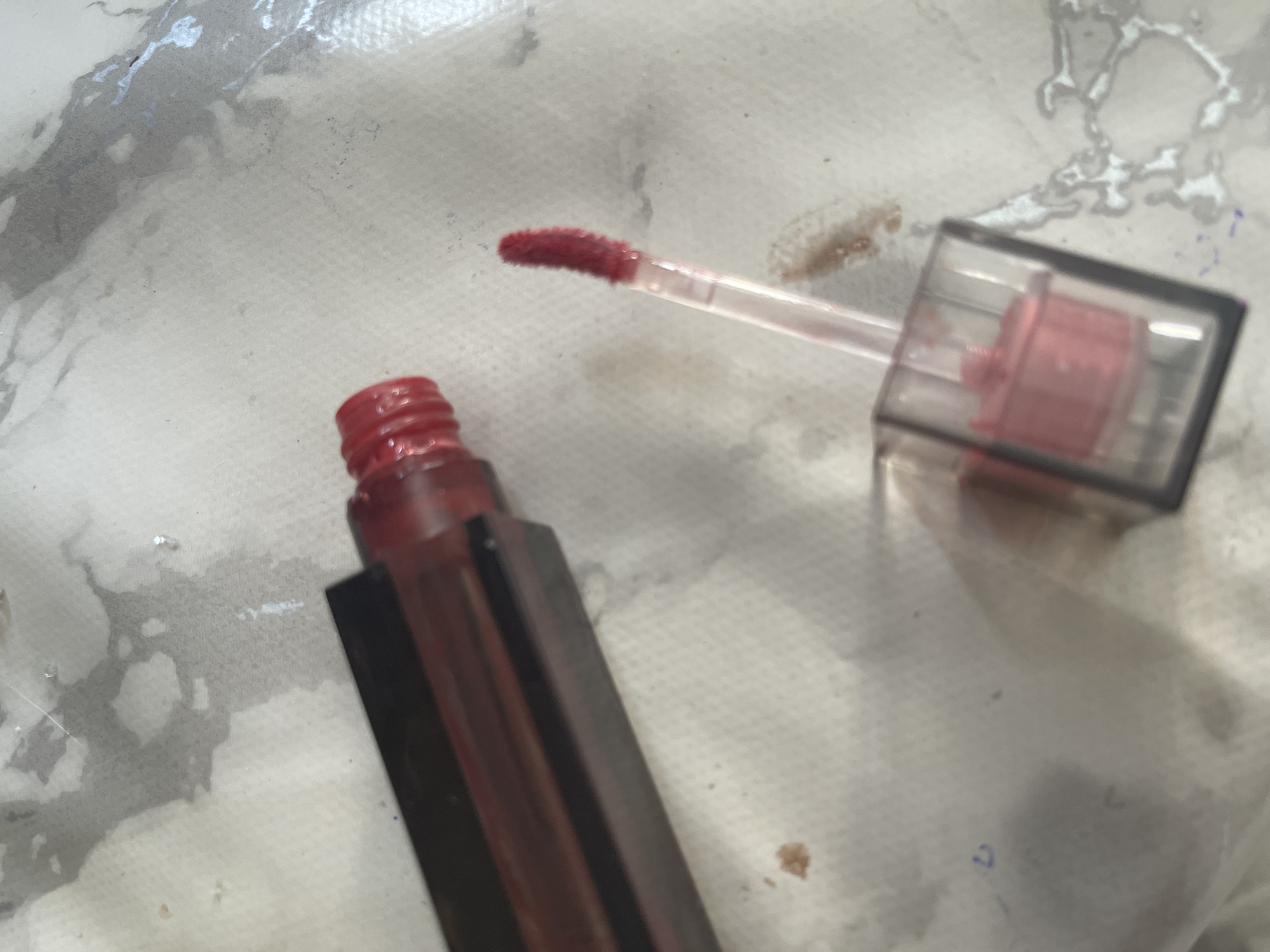 A close-up, overhead shot shows a dark brown, rectangular tube of liquid lipstick with its cap removed and lying to the left of the tube. The applicator wand, which is clear with a pink-colored liquid, is partially extended from the tube. The applicator has a fluffy, pink tip coated in lipstick. The scene appears to be on a white, textured surface with some reddish-brown smudges and water-like stains. The background is out of focus. The location in Tallinn, Estonia is not determinable from the provided image.