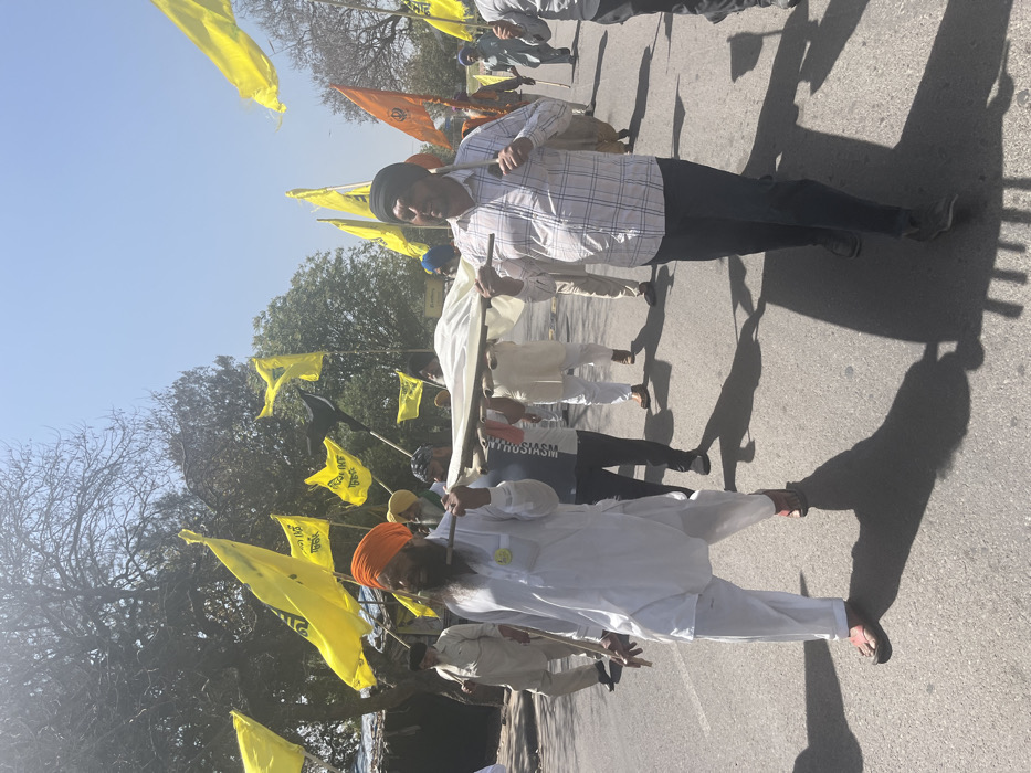 A group of men are walking down a paved street in Sahibzada Ajit Singh Nagar, India. They are carrying a white sheet on a stretcher. The men are all wearing turbans and traditional clothing. There are flags in the background. The sky is blue with some clouds.