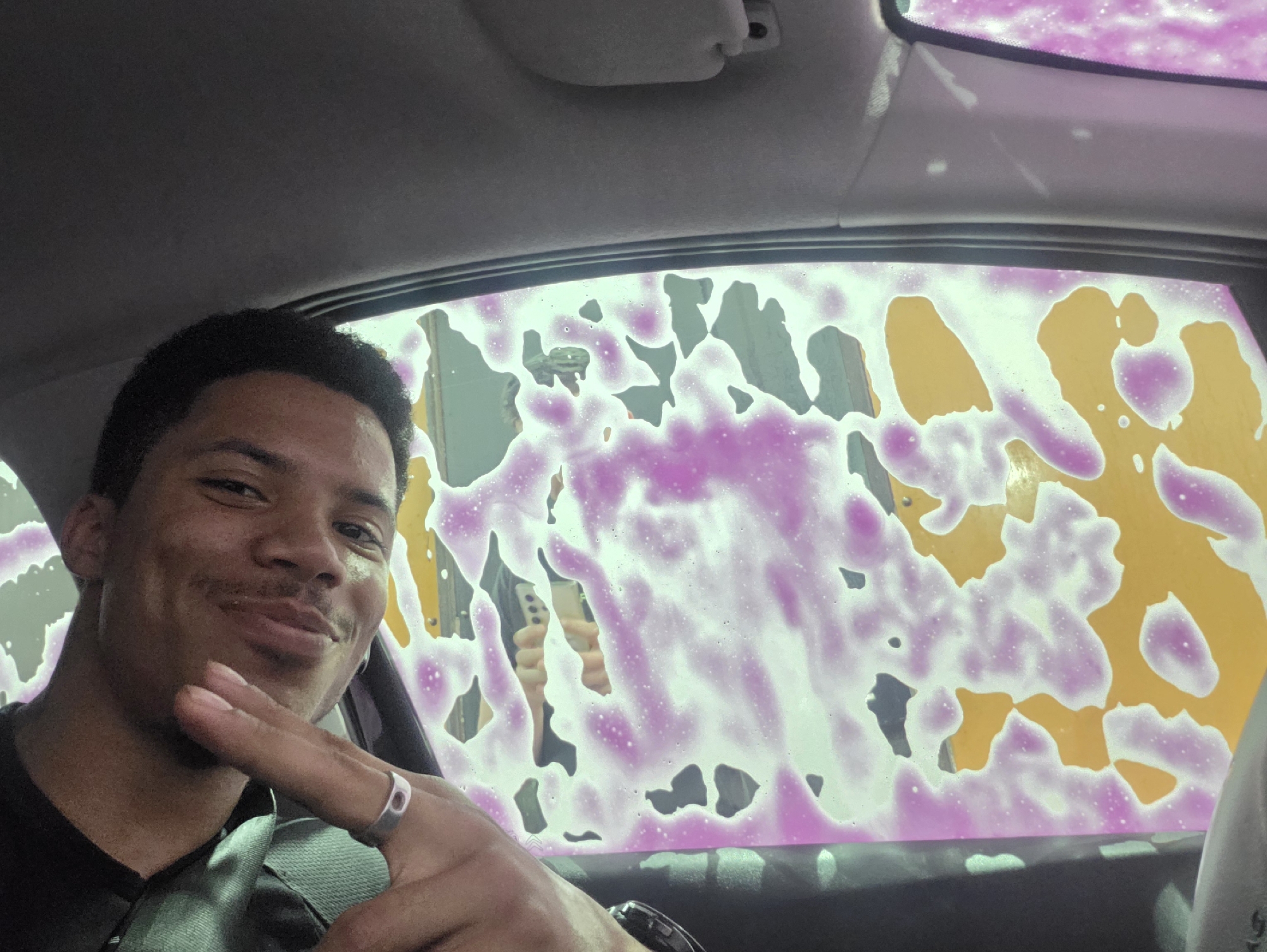 A man is inside a car, taking a selfie. He is smiling and making a peace sign with his hand. The car is being washed, and pink and yellow soap suds are visible on the windows. The background is blurred, but it appears to be a car wash. The image is likely taken in Varna, Bulgaria.