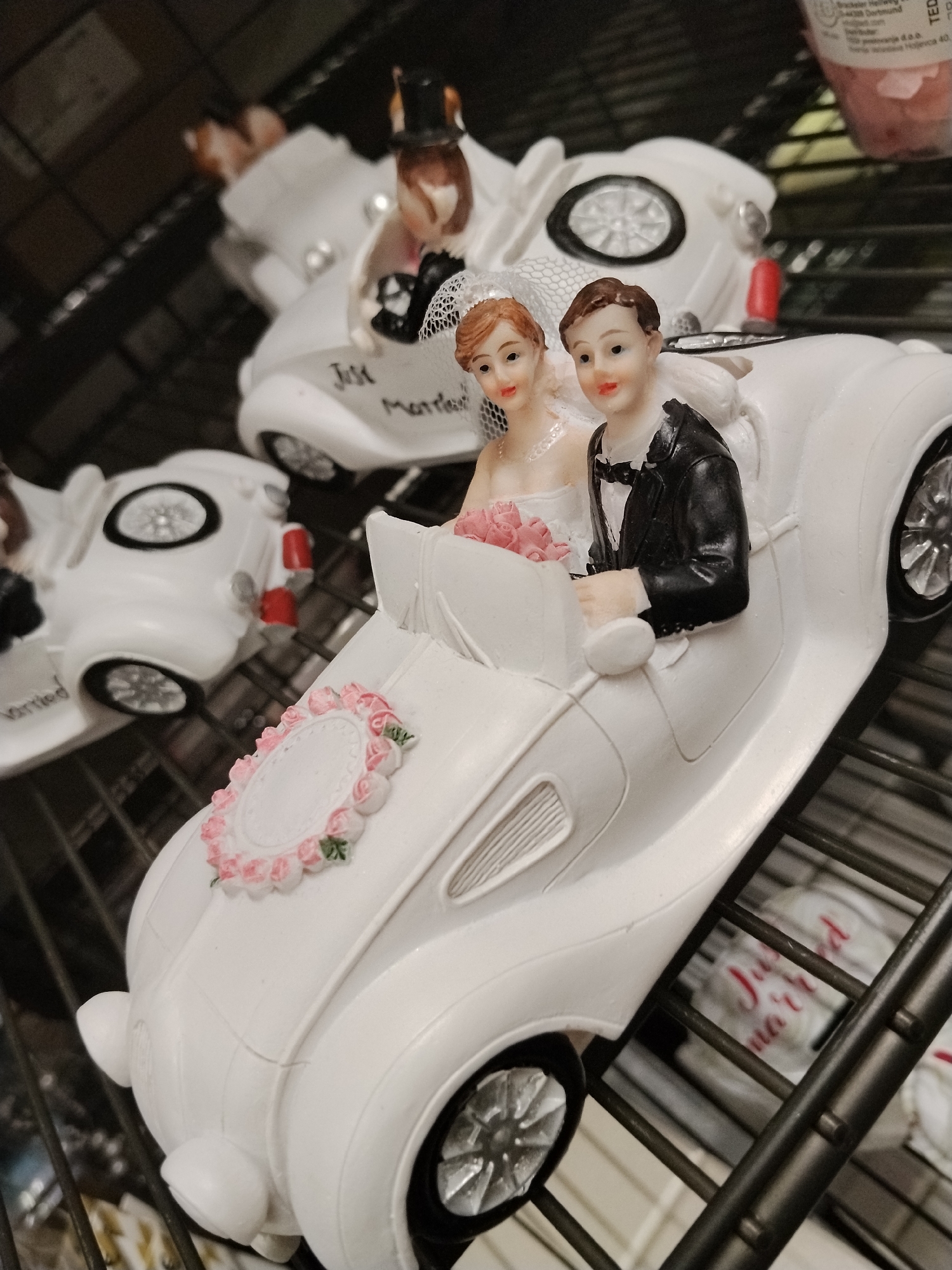 The image shows a shelf with wedding cake toppers. In the foreground, a white miniature convertible is depicted with two figurines of a bride and groom. The bride is on the left, wearing a white dress and a veil, and holding a bouquet of pink flowers. The groom is on the right, wearing a black suit and a bow tie. The car has a decorative wreath of pink roses and greenery on the hood. The text "Just Married" is visible on the side of a similar car in the background, also adorned with a bride and groom. The setting appears to be a retail environment with metal shelving. No specific location in the city of Bari, Italy, can be determined from the provided image.
