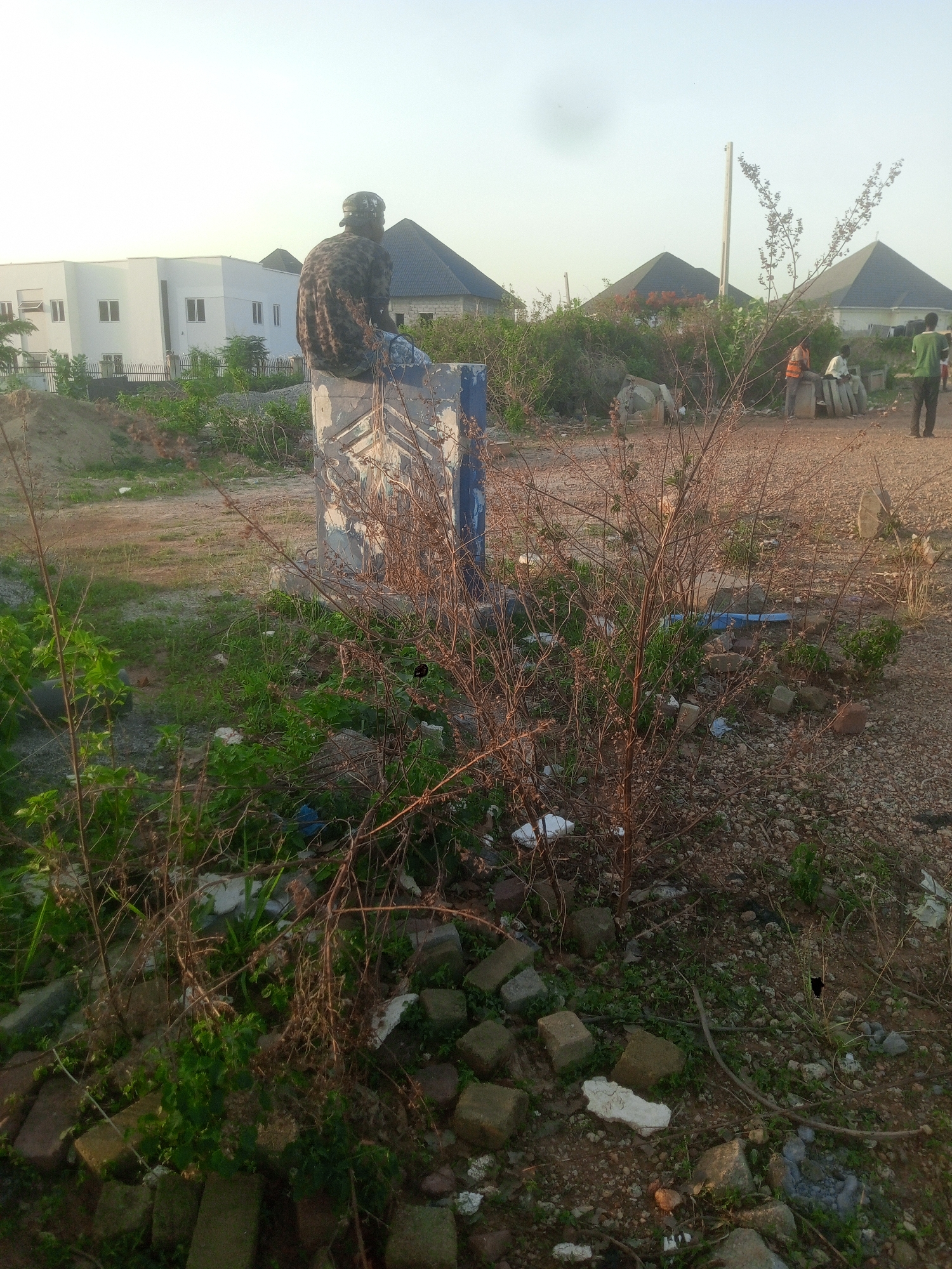 This outdoor photograph, taken in Gwarinpa, Nigeria, captures a semi-developed landscape under a bright, clear sky, likely during the late afternoon or early morning.
In the foreground, a person wearing a patterned shirt and a cap sits on a faded concrete block, facing away from the camera. The block itself shows signs of wear and has faint, geometric patterns. The immediate ground around the block is covered with reddish-brown dirt, sparse green grass, and dry, thorny bushes, along with scattered construction debris and litter like plastic bags.
In the background, a cluster of modern white buildings with dark, pyramid-shaped roofs are visible, indicating new residential development. To the right of the frame, in the mid-ground, a small group of at least three people are gathered. One individual is clearly wearing an orange safety vest, suggesting they might be workers engaged in construction or site management activities. The group appears to be conversing or resting among additional concrete slabs and debris.
The overall scene depicts an area in transition, where natural terrain meets ongoing urban expansion. The clear sky and soft lighting suggest a calm atmosphere. There is no discernible text visible in the image.