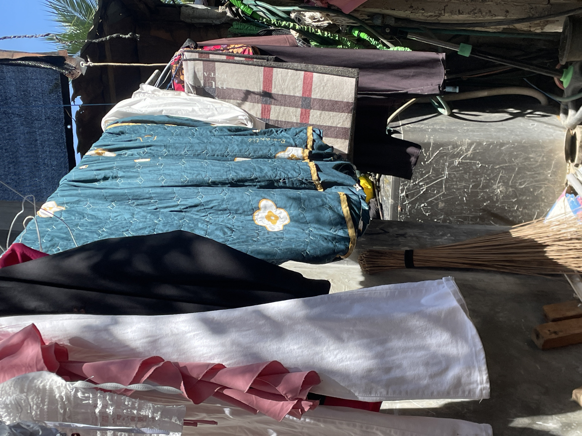 This image depicts a cluttered outdoor scene, likely a rooftop or an elevated open-air area, under bright sunlight in Davao City, Philippines. The composition is dominated by various textiles and household items, suggesting a functional domestic space.
In the mid-ground, a dark teal quilted blanket with a white floral design and the word "Romantiq" embroidered on it is prominently displayed. Below it, a stack of fabrics includes a crumpled white sheet, a smooth black fabric, and a pink fabric with ruffled edges. To the right, a traditional straw broom, bound with a black band, rests on a concrete or paved surface, next to a couple of small wooden blocks.
Above the teal blanket, a rectangular item with a beige and red plaid pattern, possibly a suitcase or a folded blanket, is visible. Partial text "TA" is discernible on its surface. The background behind these items is a jumble of wires, rods, and various colored fabrics, including greens and other shades, indicative of a storage area or a makeshift structure. Parts of what appear to be corrugated metal or plastic roofing are also present.
In the upper left corner, vibrant green palm fronds are silhouetted against a clear, bright blue sky. A rope or wire with a metal hook hangs across the upper left, suggesting a clothesline or a securing mechanism. The strong, direct sunlight creates sharp shadows and highlights the textures of the fabrics and other objects. There are no people visible, but the arrangement of items implies everyday activities like drying laundry or general outdoor storage.