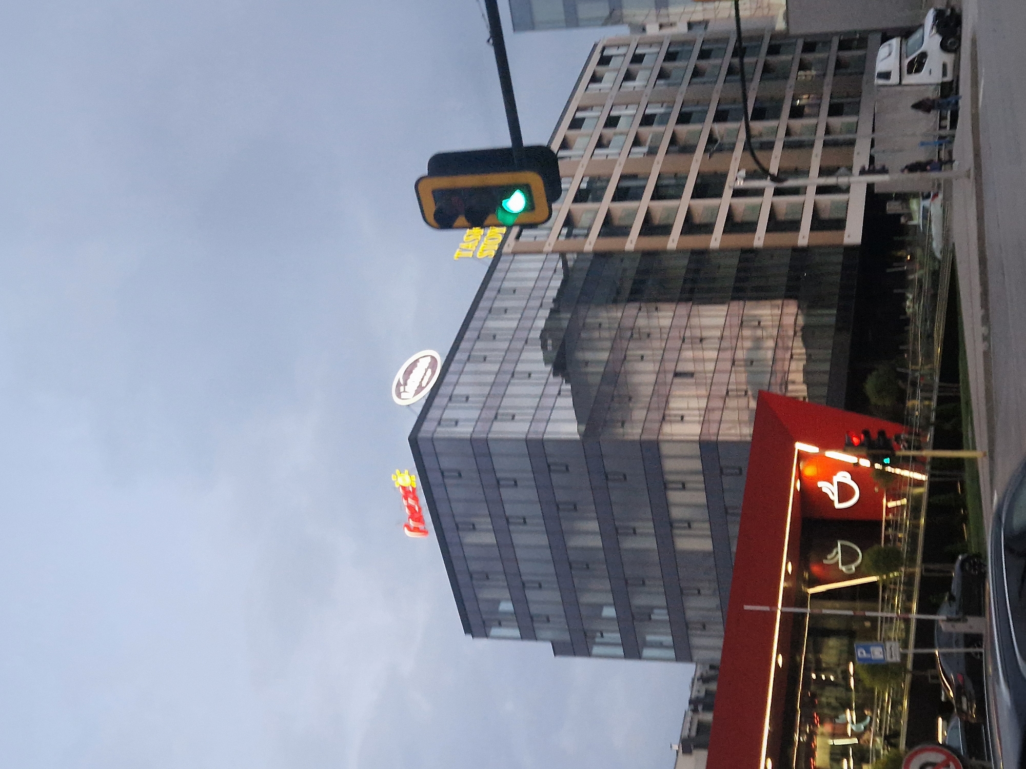 The image depicts a street scene in Unknown, Unknown, featuring a mix of architectural styles and commercial establishments. Dominating the view are several high-rise buildings, one with a glass facade reflecting the sky and another with a more traditional grid-like exterior. A prominent traffic light shows a green signal, suggesting a flow of traffic. There are recognizable commercial signs: a logo for a coffee shop and a sign that reads "Show". A red building with a coffee-related sign is situated at the lower right of the frame, possibly a cafe or drive-through coffee shop. Several cars and other vehicles are visible on the street, indicating urban activity. The sky is overcast.