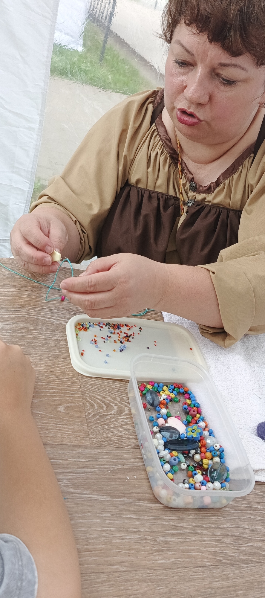 The image shows a close-up of a woman's hands as she is stringing beads. She is wearing a tan and brown tunic with decorative trim. In front of her are two containers of beads, one white and shallow with smaller, colorful beads, and a clear plastic container with larger, more varied beads. A light blue string is visible, with beads being threaded onto it. Another person's arm, wearing a grey shirt, is visible in the lower left of the frame. The scene appears to be taking place outdoors, possibly under a tent or awning, as indicated by the translucent, wrinkled material in the background, which lets in diffused natural light. The surface in front of the subjects is a wooden table. The setting does not provide specific clues to pinpoint a location within the city of Chaikovsky, Russia.