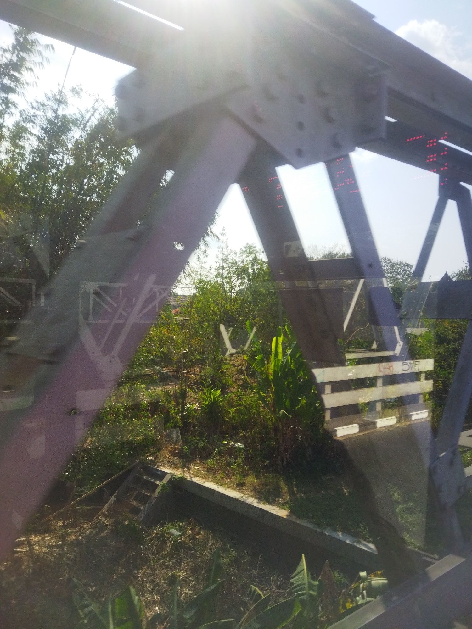 The image shows a view through a window of a train crossing a bridge in Nguter, Indonesia. The bridge is made of dark metal beams and has a railing made of white planks. The surrounding environment is lush and green with trees and other vegetation. In the distance, a few buildings are visible. The sky is blue with a few clouds. The sun is shining brightly, creating lens flare in the image. The train tracks are visible on the bridge, and there are some red lights on the bridge structure. The overall impression is of a sunny day in a rural area.