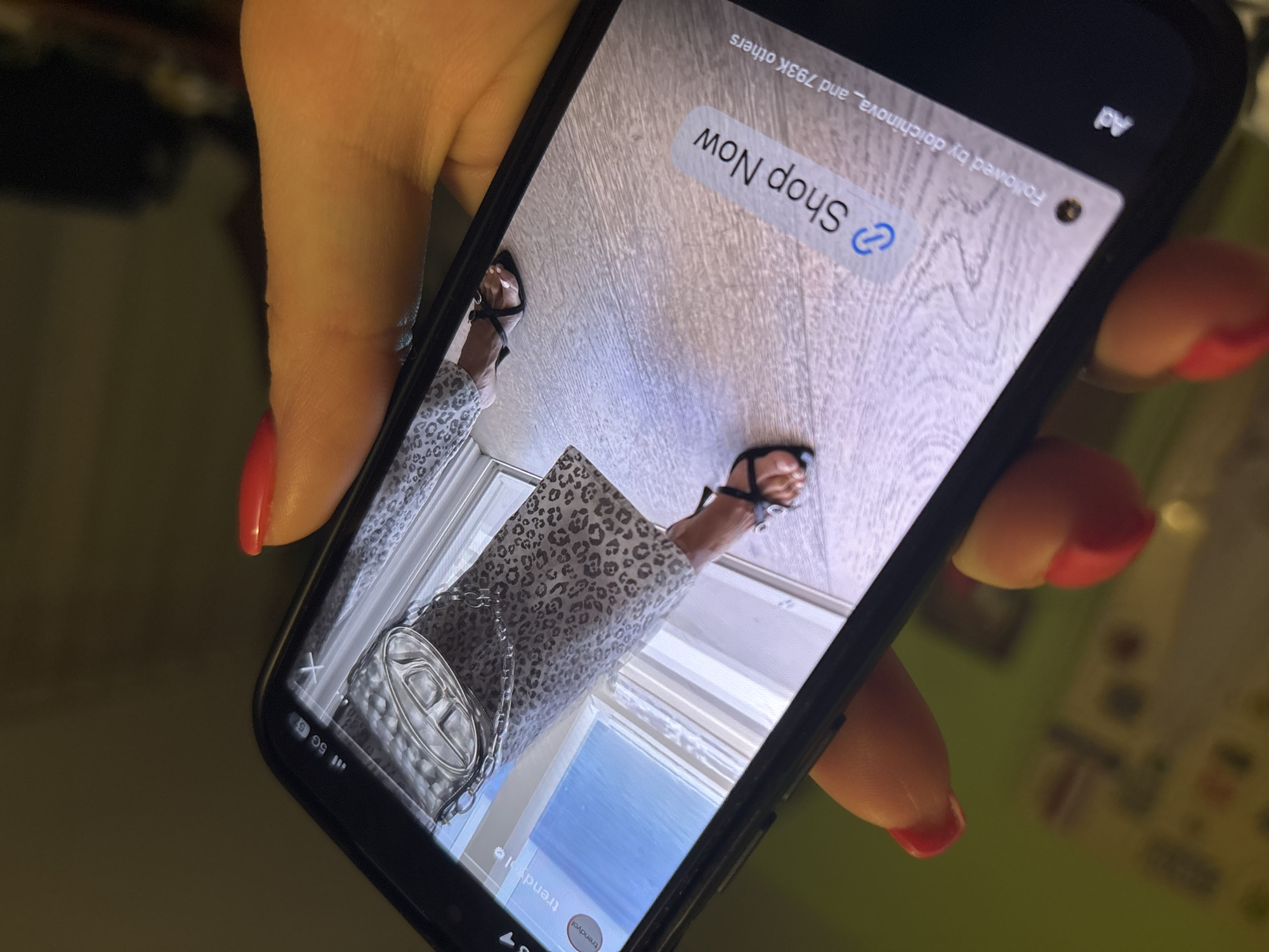 A photograph shows a person's hand holding a smartphone. The smartphone screen displays an advertisement for "Shop Now" featuring a model standing on a light-colored wooden floor wearing a leopard-print outfit, black sandals, and carrying a silver handbag. The lower portion of a window is visible in the reflection of the handbag. The hand holding the phone has red nail polish. The background is blurry and shows hints of what could be a domestic interior. The location is not discernible from the given image. There is no indication that the photo was taken in Sofia, Bulgaria.