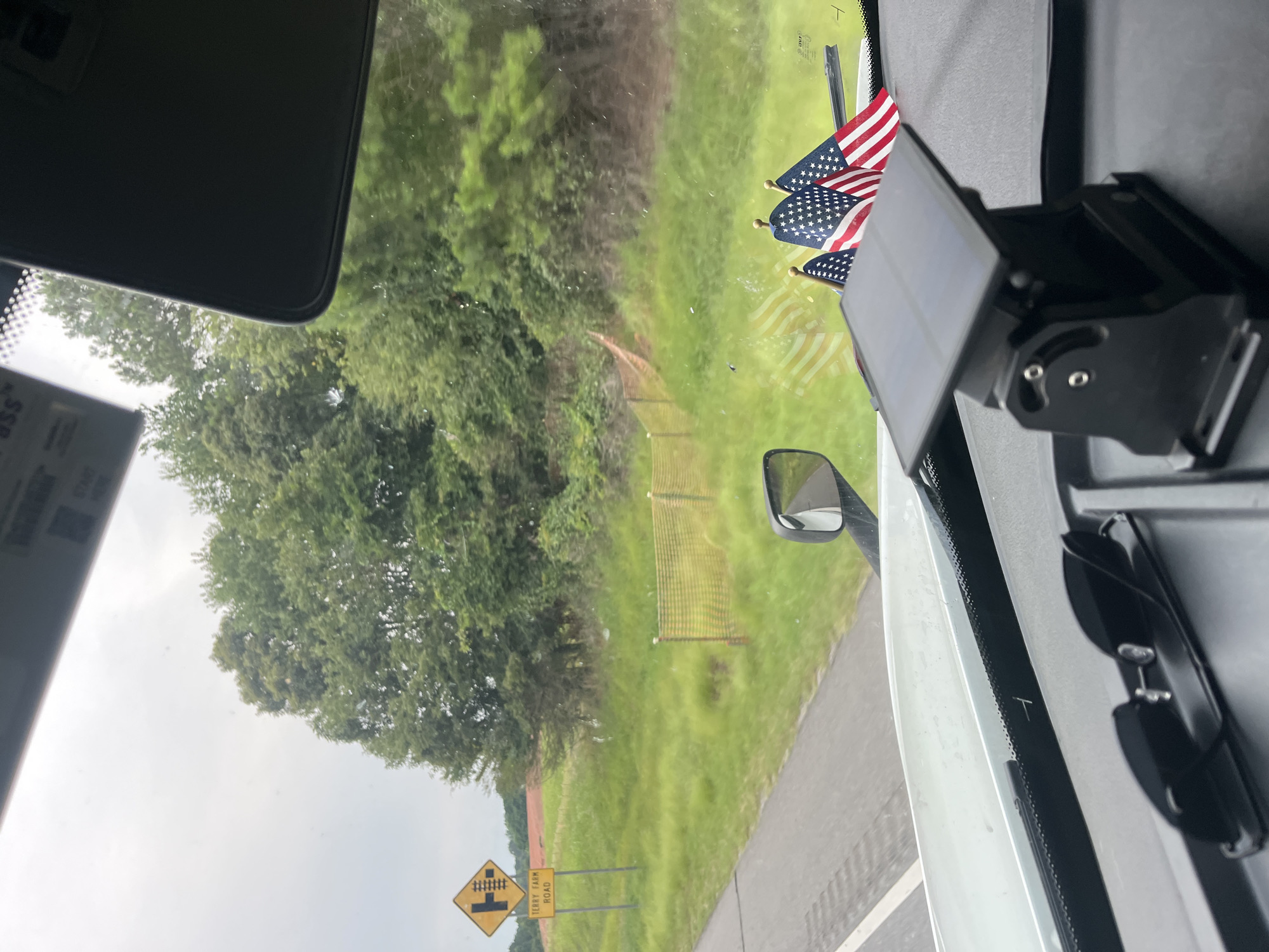The image shows a view from inside a vehicle, likely a truck, looking out the windshield. In the foreground, on the dashboard, are a pair of black sunglasses and a black solar-powered device with an adjustable mount. Behind these objects, three small American flags are mounted on poles.
Outside the vehicle, the landscape consists of a grassy area with trees in the background. A yellow diamond-shaped road sign with black symbols indicating a railroad crossing and a road intersection is visible on the left. Below this sign is another rectangular yellow sign reading "TERRY FARM ROAD". A section of orange safety fencing is also present in the grassy area.
The sky is overcast, suggesting a cloudy day. The vehicle appears to be on a road, with a portion of the road visible to the left of the image. There are no people visible in the image. The overall scene depicts a roadside view from a vehicle, possibly in a rural or semi-rural area. The location is identified as Arcade, United States of America, based on the sign reading "TERRY FARM ROAD".