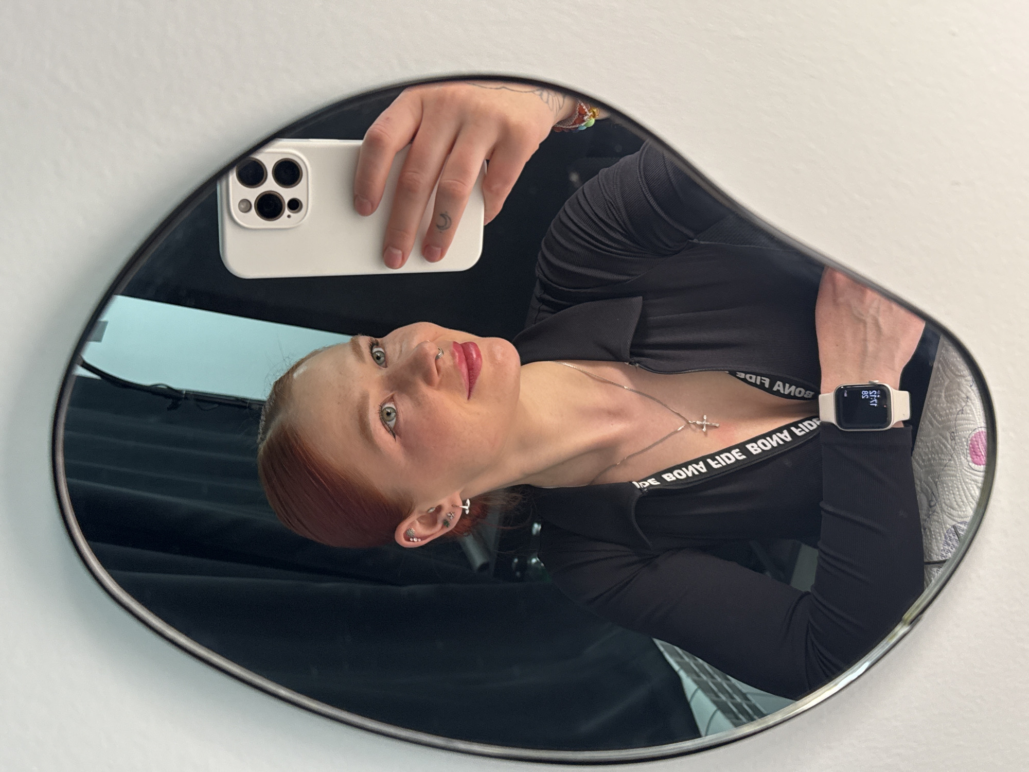 The image shows a mirror selfie of a female individual with reddish-brown hair, captured from the chest up. She is looking to her right, displaying a slight smile. Her eyes are light blue/green, and she wears light makeup, including eyeliner and pink lip gloss. A silver-colored septum ring is present, along with multiple piercings in her left ear, featuring various studs and hoops. She wears a black, long-sleeved, ribbed top with a collar and a zippered front. A silver-colored chain with a cross pendant is visible around her neck. On her left wrist, she wears a white smartwatch displaying inverted text and numbers, including "59," "31:37," and "65." Her right hand holds a white smartphone with a visible multi-lens camera array, utilized for capturing the reflection. A small crescent moon tattoo is present on the middle finger of her right hand, and a multi-colored beaded bracelet is partially visible on her right wrist. The mirror itself is irregularly shaped, with a thin dark frame, and is mounted on a white wall. The background reflected in the mirror behind the individual includes dark, vertically aligned drapes and a segment of a light blue or turquoise surface. The individual's left arm rests on a white, textured paper towel or disposable covering, with a portion of a white radiator or similar fixture visible beneath it. The specific location within Rostov-on-Don, Russia, cannot be determined from the visual content of the image.