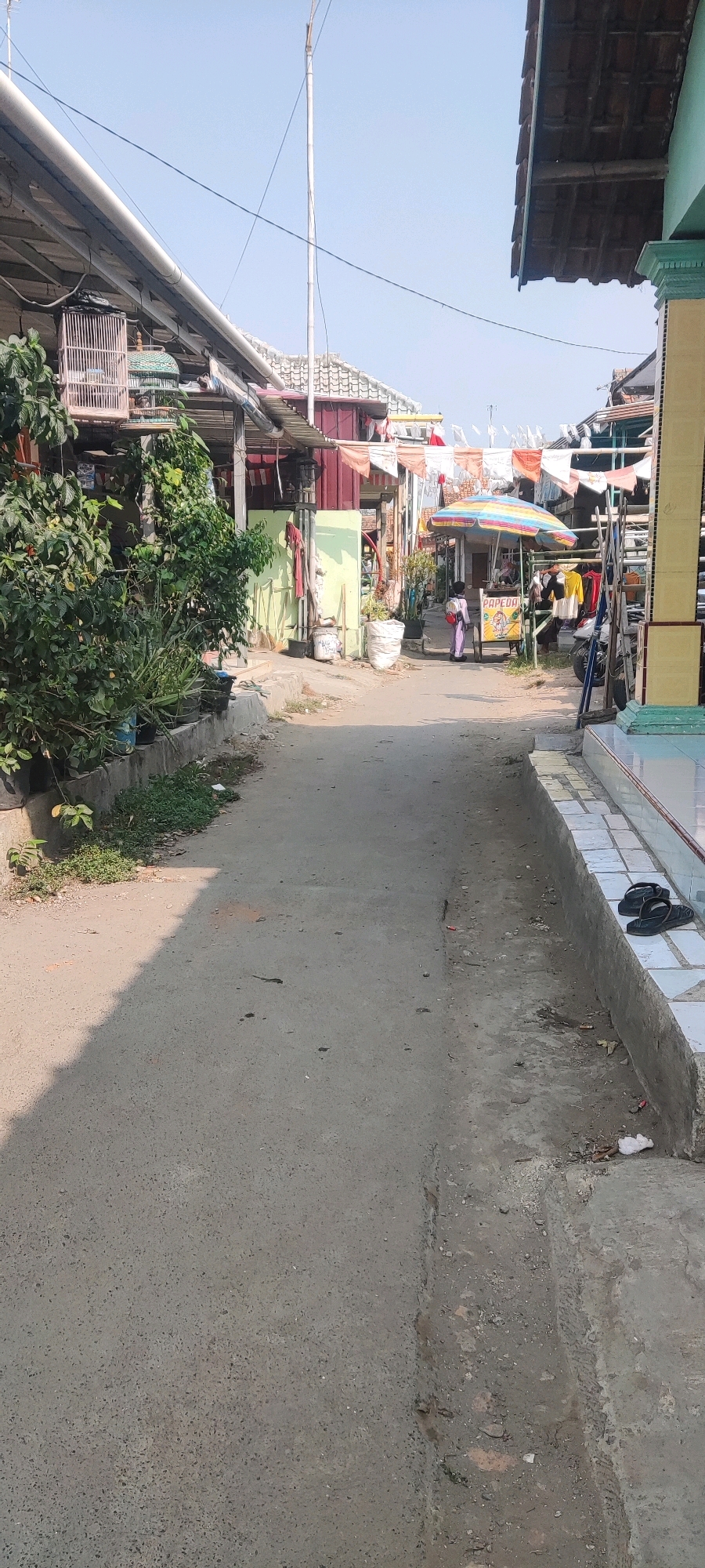 The image depicts a narrow paved pathway in Gebang, Indonesia, lined with buildings on either side. On the left, a dense green bush is prominent, with potted plants below it and a birdcage suspended from a structure above. The pathway itself is a muted gray, with some debris and a shadow cast from the left. On the right, the pathway is bordered by a raised concrete structure with white tiles, on which a pair of black flip-flops rests. Further down the path, a child wearing a pink outfit walks away from the camera towards a food stall that has a colorful umbrella and a cart displaying a sign with the word "PAPEDA." Various items, including clothing and a motorcycle, are visible near the stall. Overhead, laundry hangs on a line, and a tall, thin pole extends vertically into the clear blue sky. The overall scene suggests a residential or commercial area with everyday activities taking place.