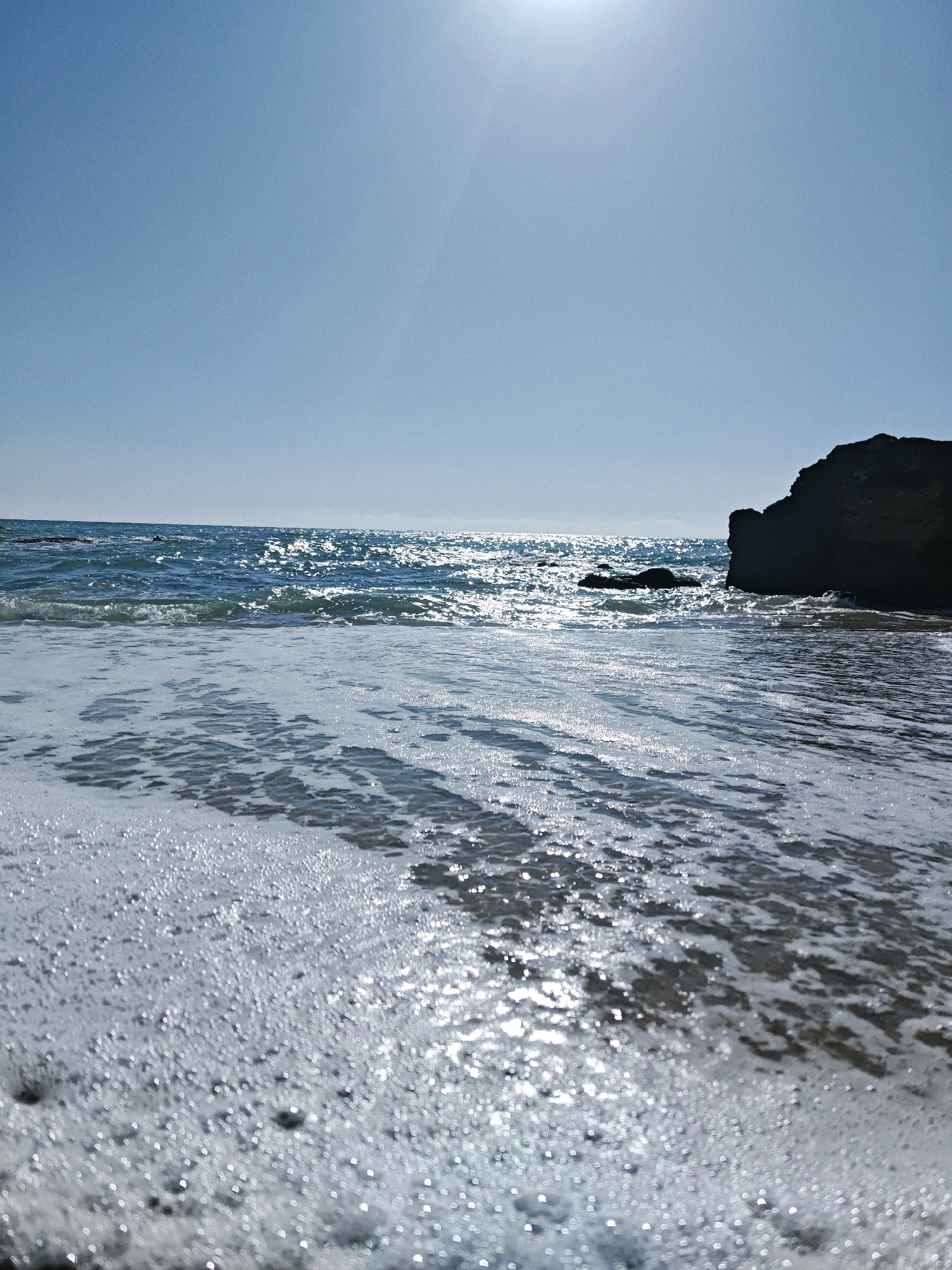 This is a low-angle, eye-level, wide shot of a sunny beach with the camera focused on the water and the foam of the waves as they recede from the shore. The sky is a clear, bright blue with the sun high and to the left, casting a bright glare on the water. The ocean is a deep blue with whitecaps and sunlight glinting off the surface. To the right, a large, dark rock formation juts out of the water. The foreground is dominated by a carpet of white foam and bubbles as a wave washes ashore. The overall impression is of a bright, peaceful, and refreshing day at the beach.
