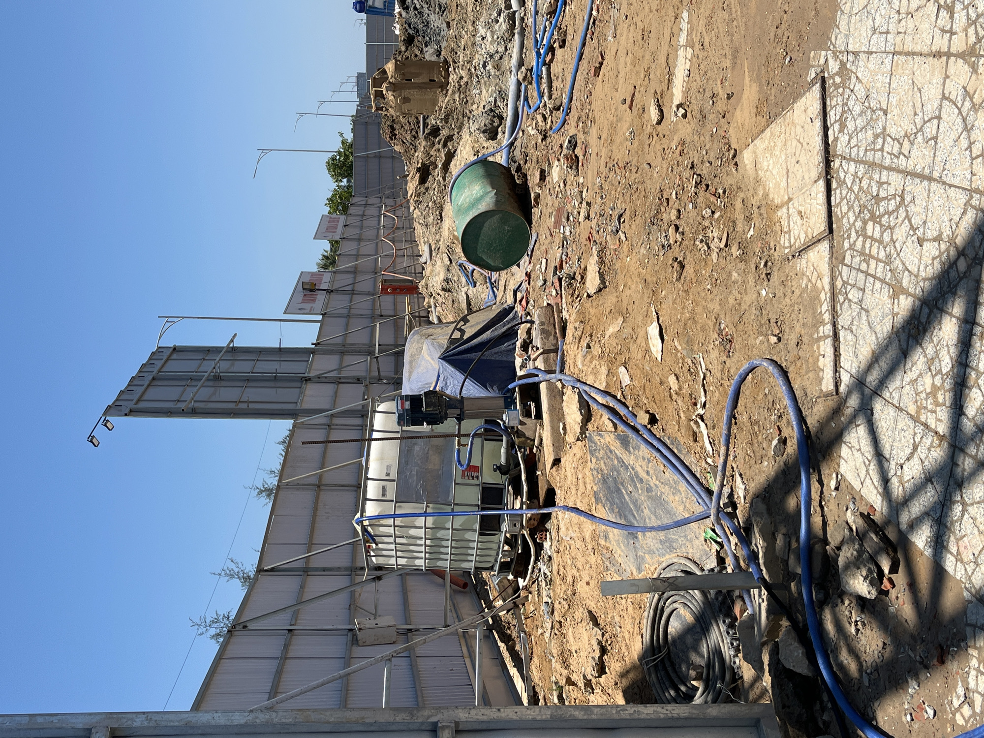 The image depicts a construction site in an urban area, possibly Đà Nẵng, Vietnam, based on general architectural features and the environment. The foreground shows a dirt and rubble ground. In the center, there is a large white plastic water tank with a metal cage around it, partially covered by a blue tarp. A green metal drum sits near the tank. Blue water hoses are scattered on the ground, connecting to the tank and running off-frame. To the left, a metal fence surrounds the construction site with billboards and streetlights visible in the background. The background also contains piles of construction materials and dirt. The sky is clear and blue. Some tiled ground is on the right side.