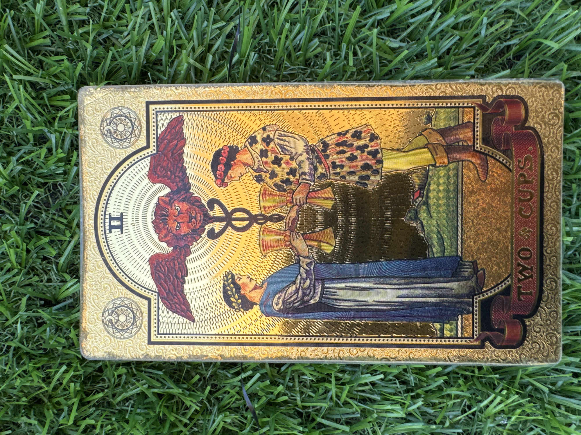 The image shows a tarot card, specifically the "Two of Cups" card from a golden-colored deck, lying on a patch of artificial grass. The card features two figures in period clothing holding cups towards each other, with a winged lion-like creature with a caduceus above them. The card is bordered in gold and has ornate decorative elements. The background shows a golden color. The card is lying horizontally on the grass. There are no identifiable elements related to Đà Nẵng, Vietnam.