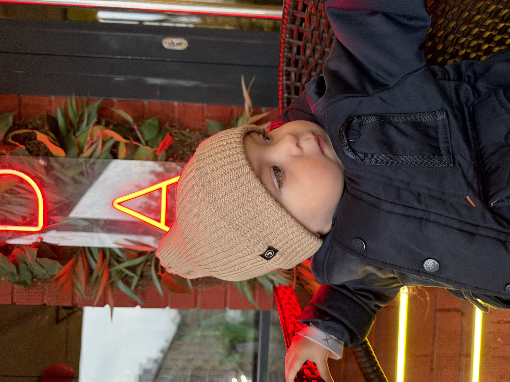 An infant, approximately one year of age, is seated in a dark brown wicker chair within what appears to be a commercial establishment in the city of Votkinsk, Russia. The child wears a light tan ribbed knit beanie with a black square patch featuring a circular white logo on its left side. A dark blue or black quilted jacket with large dark buttons and an orange stitching detail on the lower-right side covers the child's torso. A black fabric loop is visible on the jacket's upper-right chest. The child's left hand grasps a horizontal red illuminated bar, and a white cuff is visible at the wrist. The child's gaze is directed towards the right side of the frame.
The background features a prominent red brick wall adorned with artificial green and reddish foliage. A transparent panel, likely glass, displays two neon signs: a partial red "D" in the upper center and a prominent yellow-orange triangular "A" shape without a crossbar, located to the right of the center. To the left of the child, a large window reveals a blurry urban exterior with overcast lighting and distant building structures. In the upper-left corner, a spherical red object is partially visible. To the right of the child, a dark vertical structure, possibly a door frame or column, features a rectangular recessed handle. A linear yellow-orange light source is positioned horizontally in the lower-left foreground.