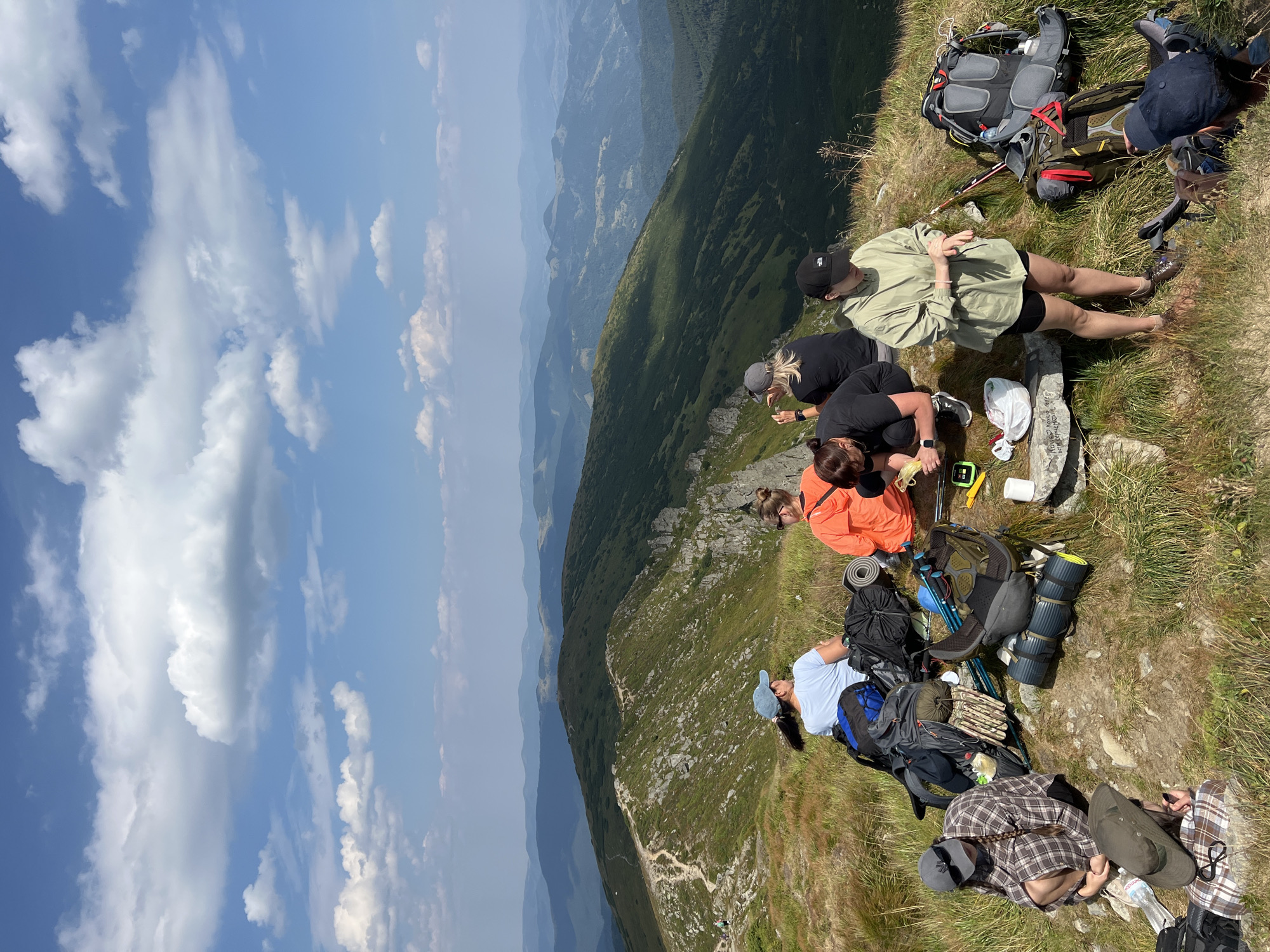 The image depicts a group of hikers taking a break on a mountain ridge in Vorokhta, Ukraine. The landscape is characterized by rolling green hills and mountains under a bright blue sky with scattered white clouds. A well-worn hiking path is visible snaking along the ridge in the background. In the foreground, several individuals are seated and crouching amongst their camping gear, which includes backpacks, sleeping mats, and walking sticks. One person is seen eating, while others appear to be resting or conversing. The overall scene conveys a sense of outdoor recreation and camaraderie amidst a scenic natural environment.