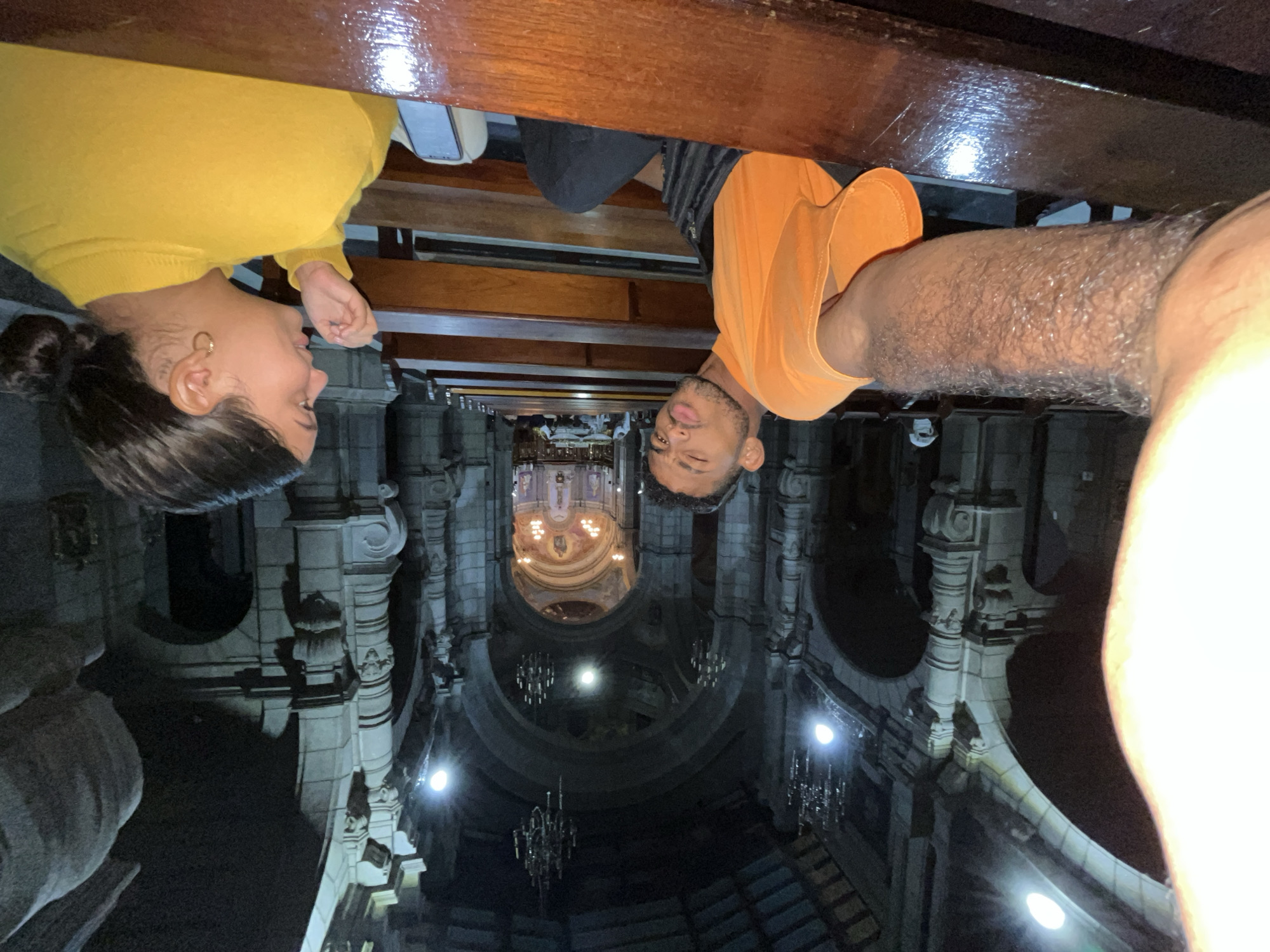The image depicts two individuals, a man and a woman, seated on wooden pews inside a large, dimly lit church, likely the Basilica Menor de Nuestra Señora de la Inmaculada Concepción in Mérida, Venezuela. The man, wearing an orange t-shirt, is in the foreground, taking a selfie and making a kissing face. He has a black bag slung across his shoulder. The woman, wearing a yellow sweater, is seated to his right, looking towards the altar in the distance. Her hair is pulled back in a bun, and she wears hoop earrings. The church's architecture features high vaulted ceilings, ornate stonework, and chandeliers, with the altar area brightly lit in the background. The lighting is low, creating shadows and highlighting the architectural features of the church. The pews are made of dark polished wood.