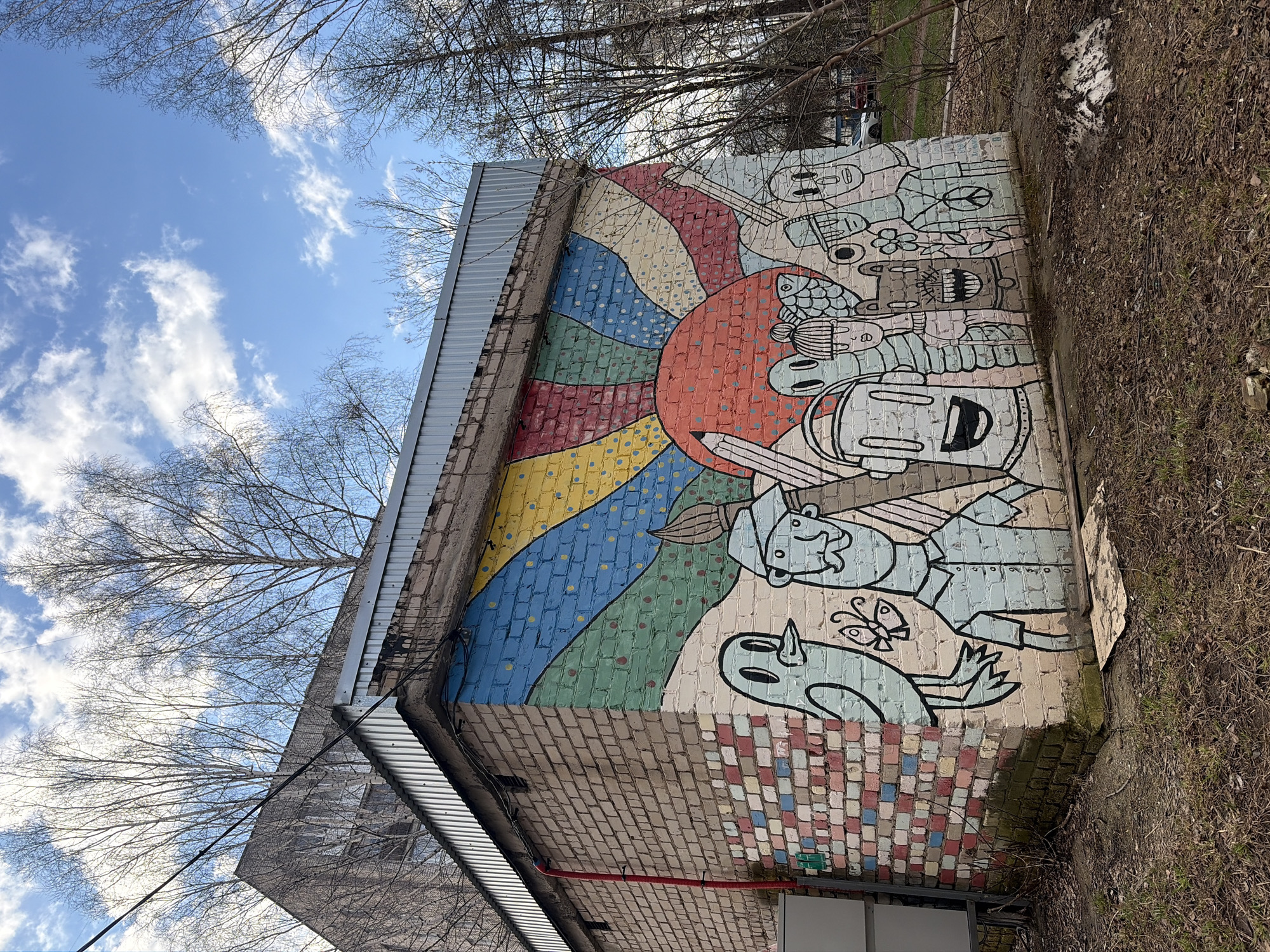 The image showcases a vibrant and whimsical mural painted on the brick wall of a building in Chaikovsky, Russia. The scene is captured on a clear, sunny day, likely in early spring, indicated by the bare branches of deciduous trees and patches of dry earth and melting snow on the ground.
The mural itself features a diverse array of cartoon-like characters and a colorful abstract background. A large, semi-circular design, resembling a sun or rainbow, spans the upper left portion of the wall, with segments of red, orange, yellow, green, blue, and purple, some dotted with contrasting colors.
In the foreground of the mural, a central, blocky character with a helmet-like head holds what appears to be a paintbrush or sword. Its face is marked with a prominent, stylized 'D' or a wide, simple smile. To its left, another creature, possibly a bird or frog, is depicted with a surprised expression, and a butterfly hovers nearby. Above and to the right of these figures, a medley of smaller, quirky characters fill the space, including a fish, a guitar-like object, and various human-like and abstract forms with unique expressions. All characters are outlined in black and subtly filled with pastel shades of blue, pink, gray, and green.
The building's wall is made of light-colored bricks, some unpainted below the mural, and it features a corrugated metal roof or awning along its upper edge. The surrounding environment includes a bright blue sky dotted with white clouds. Bare trees with intricate branch networks frame the scene. The ground adjacent to the wall consists of brown earth, dry leaves, and sparse grass, with a small patch of melting snow or mud visible to the far right. Faint outlines of vehicles can be seen through the trees in the background. The mural's playful artistry provides a striking contrast to the utilitarian architecture and early spring landscape. No explicit text is legible within the mural.