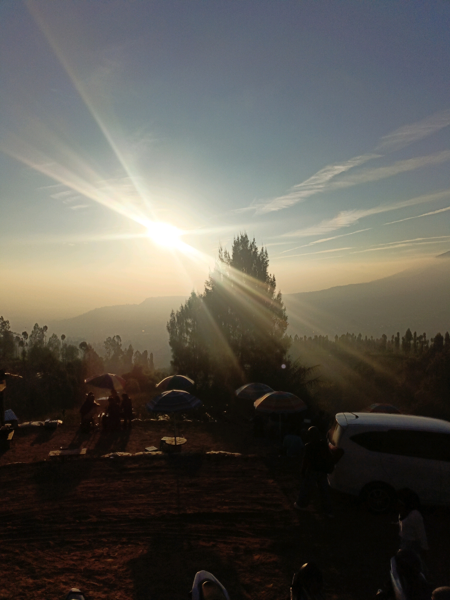 The image captures a serene outdoor scene at sunrise in Butuh Kidul, Indonesia, bathed in the warm glow of the ascending sun. The sky is a gradient of pale blue, transitioning into a soft yellow and orange hue near the horizon, with thin, wispy clouds streaking across it. The sun itself is a prominent, bright star, emitting powerful rays that dramatically illuminate the landscape and cast long shadows.
In the foreground, a group of people are gathered, some seated and others standing, around several umbrellas that provide shade. The ground appears to be a mix of dirt and sand. A white van is parked nearby, its presence suggesting travel or a camping trip. The overall atmosphere is peaceful, with the natural beauty of the landscape enhanced by the golden light of dawn. The scene is set against a backdrop of rolling hills and dense trees, suggesting a rural or elevated location. The presence of the van and umbrellas indicates a recreational activity, possibly a picnic or a scenic viewpoint visit.