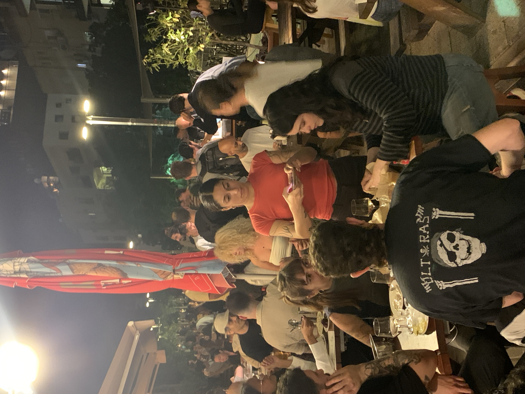 The image captures a crowded outdoor setting, likely a restaurant or bar, in the city of Tel Aviv, Israel. Many people are seated at wooden tables, engaged in conversation and activities such as drinking and eating. The lighting suggests it is nighttime, illuminated by street lamps and overhead lighting. The people are dressed in casual attire, and a variety of hairstyles and ages are represented. The presence of a large, colorful umbrella indicates an attempt to provide shade. The environment appears lively, with a notable buzz of social interaction.