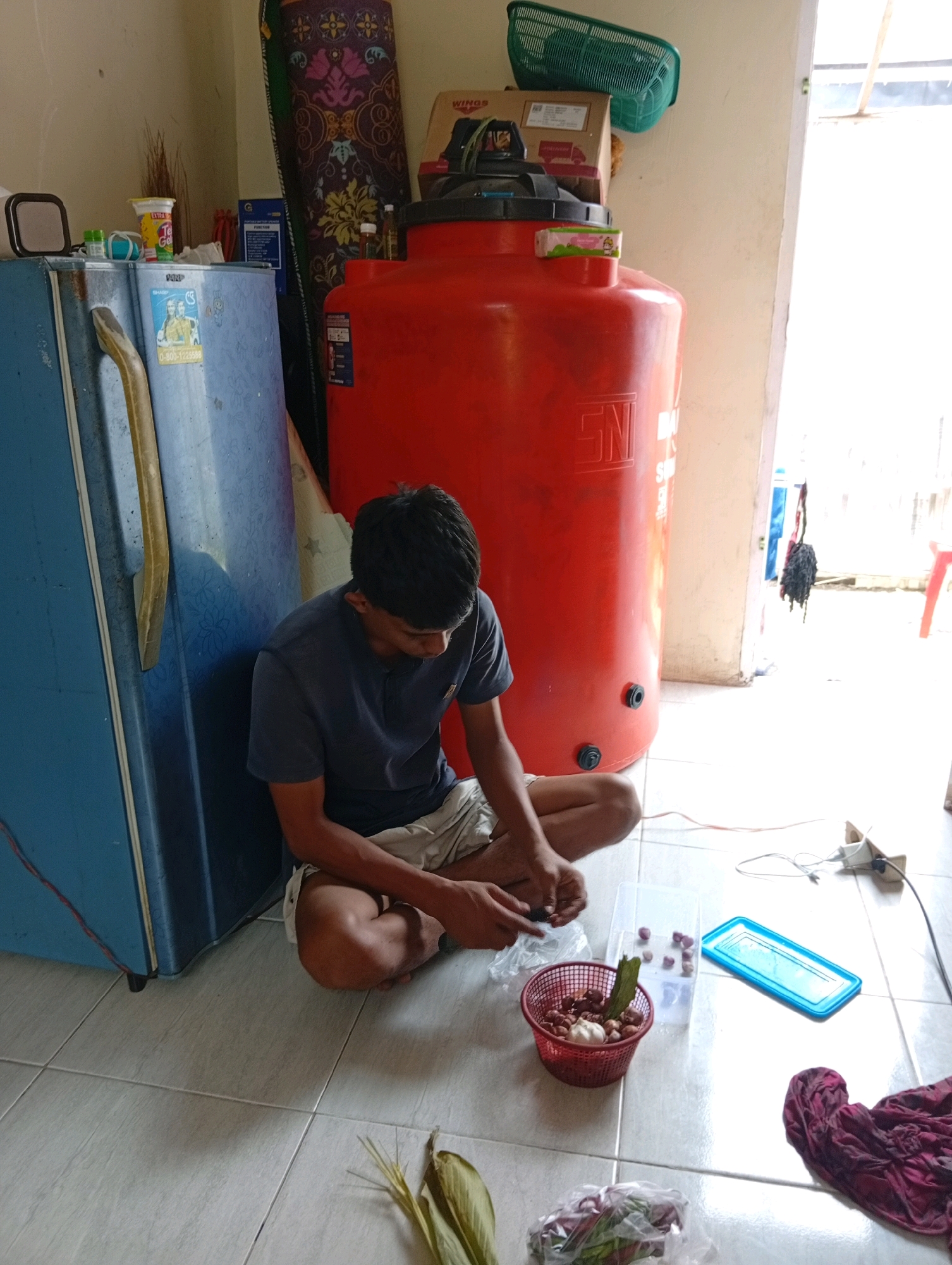 The image depicts an indoor scene, likely in Kualu, Indonesia, featuring a young man sitting on a tiled floor next to a blue refrigerator and a large orange water tank. The man is wearing a dark blue t-shirt and light-colored shorts, and he is actively engaged in preparing food, specifically peeling small onions or shallots. He holds a small knife in his right hand and appears to be working over a red basket filled with ingredients, which also contains a head of garlic and some green leaves. In front of him, there are several clear plastic containers with some of the peeled onions and a blue-lidded container. To the left of the man, a blue refrigerator stands against the wall. Behind the man and to his right is a prominent orange water tank, possibly a large household water storage unit. A piece of patterned cloth is draped behind the water tank, and a green plastic basket is visible on top of some boxes. Electrical cords and a power adapter are present on the floor near the water tank, suggesting a power outlet is nearby. The environment appears to be a functional, possibly residential, space.