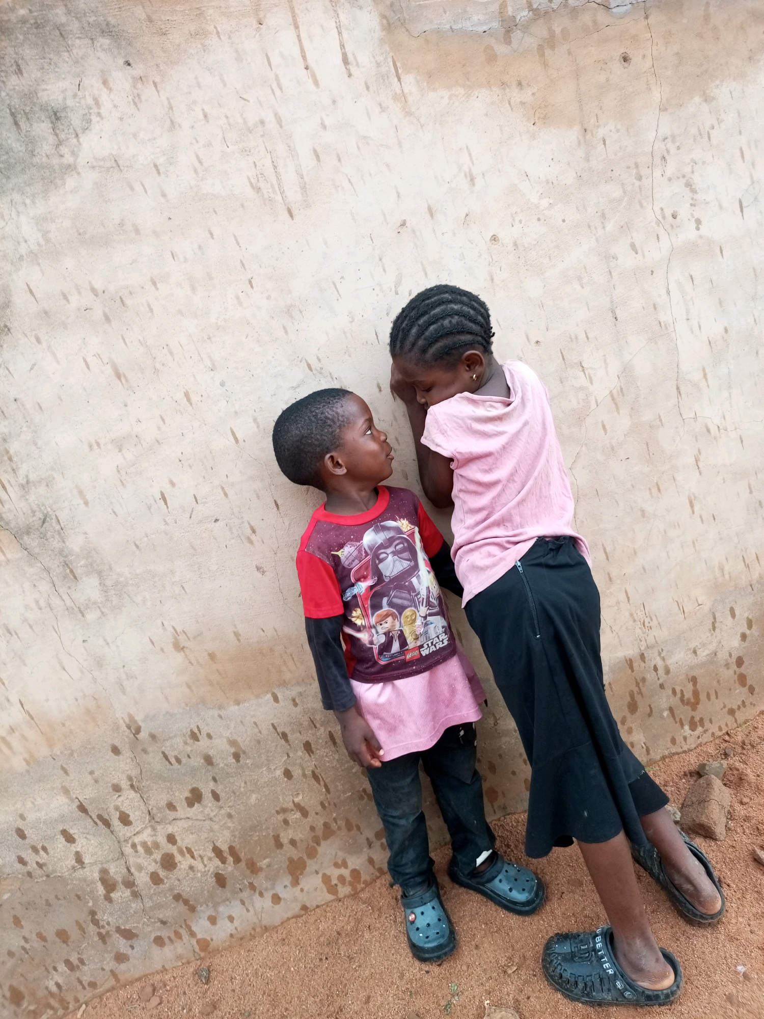 Two young children, one standing and facing left, the other leaning against a rough, beige-colored wall and facing right, appear to be in an outdoor setting. The standing child, dressed in a red long-sleeved shirt with a graphic print and dark pants, is looking up at the other child. The child leaning against the wall wears a light pink t-shirt and a black skirt. Their faces are close, suggesting an interaction or conversation. The wall has a textured surface with some stains and cracks, typical of exterior building material. The ground beneath them is sandy and reddish-brown, with a few small rocks scattered around. This scene is located in Tempure, Nigeria, outdoors.