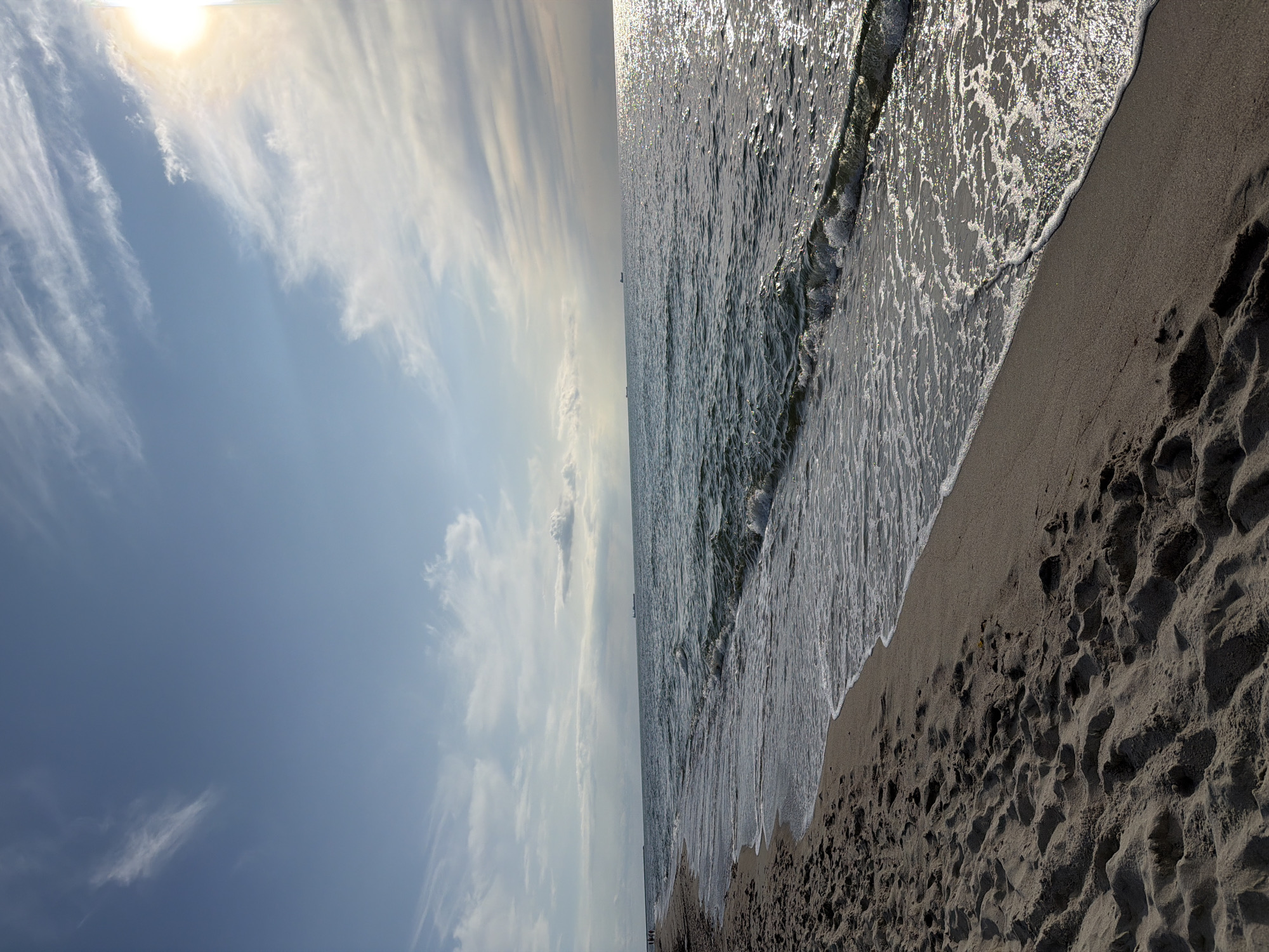 The image depicts a serene coastal scene, likely the beach in Baltiysk, Russia, on a bright, partly cloudy day. The foreground is dominated by textured sand, showing footprints, indicating recent human presence. A gentle wave is breaking on the shore, leaving a line of white foam and glistening water on the wet sand. The sea stretches to the horizon, its surface shimmering with sunlight. Distant cargo ships are visible on the water, suggesting maritime activity. The sky is a soft blue, with wispy white clouds streaked across it, and the sun is a bright, slightly diffused orb in the upper right corner, casting a warm glow and reflecting off the water. The overall atmosphere is tranquil and natural.