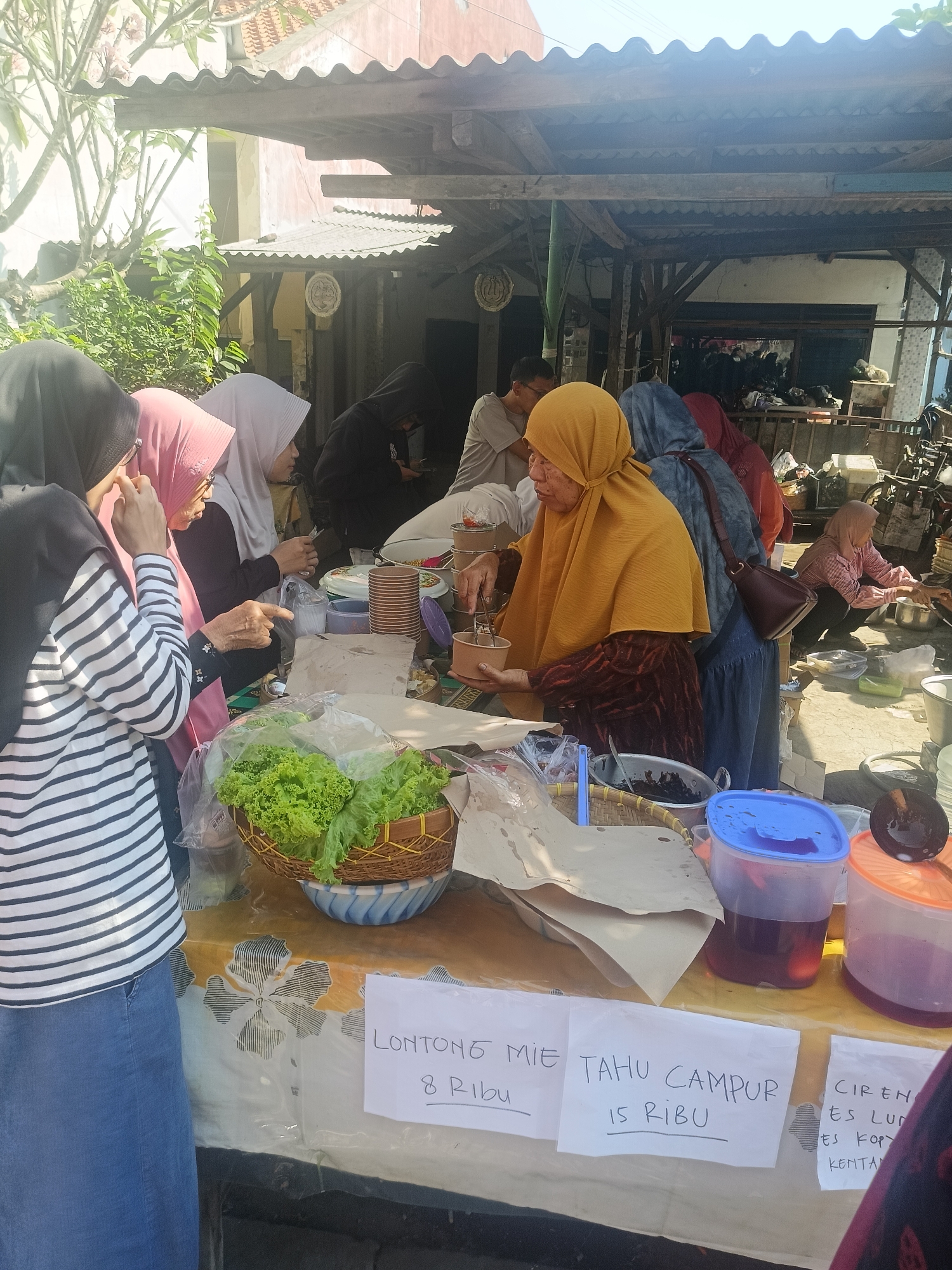 The image shows an outdoor food stall in Gedangan, Indonesia. Several people are present, mostly women wearing hijabs. A woman in a yellow hijab appears to be serving food, holding a bowl and utensil. In the foreground, there is a basket of lettuce, brown paper wrappers, and containers of drinks. Signs are visible with handwritten prices and food item names like "LONTONG MIE 8 RIBU", "TAHU CAMPUR 15 RIBU", and "CIRENG ES LU...". The stall is set up under a wooden shelter with a corrugated roof. In the background, more people are visible, and there is a cluttered area with various objects, possibly storage or discarded items. The overall scene suggests a bustling local market or street food vendor.