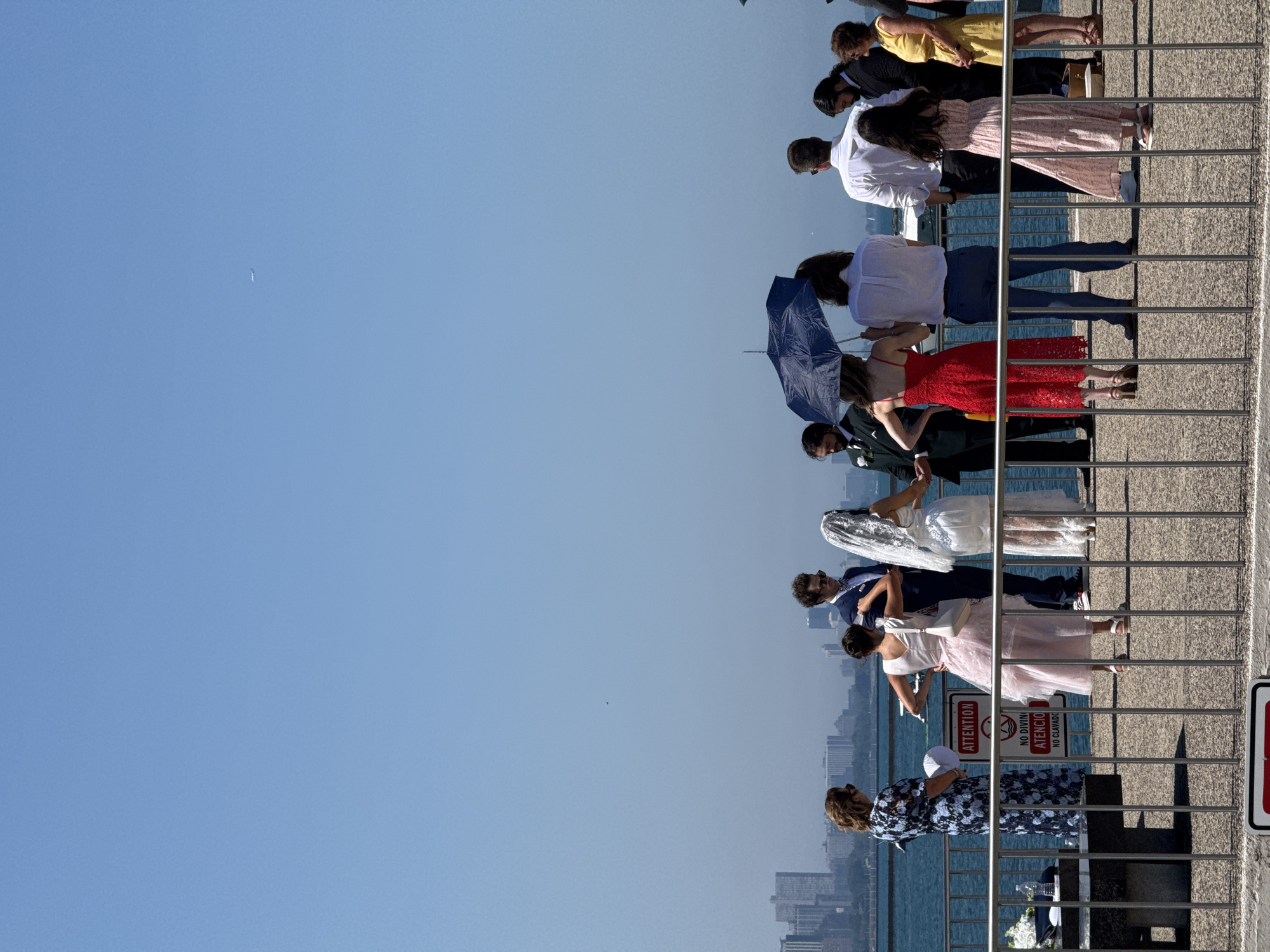 The image captures a wedding ceremony taking place outdoors on a bright, sunny day with a clear blue sky. The scene is set on a pier or boardwalk overlooking a body of water, likely Lake Michigan, with a cityscape of Chicago in the background. A group of people, dressed in formal and semi-formal attire, are gathered. At the center, a bride in a white wedding dress and veil stands next to a groom in a dark suit. Other guests, including women in colorful dresses and men in suits, surround them. One woman holds a dark umbrella, suggesting it might be for shade rather than rain. To the left, a woman in a patterned dress stands near a table with some wedding arrangements. A sign with "ATTENTION" and "NO DIVING" in English and Spanish is visible on a railing. The overall atmosphere appears to be that of a casual, intimate wedding celebration.