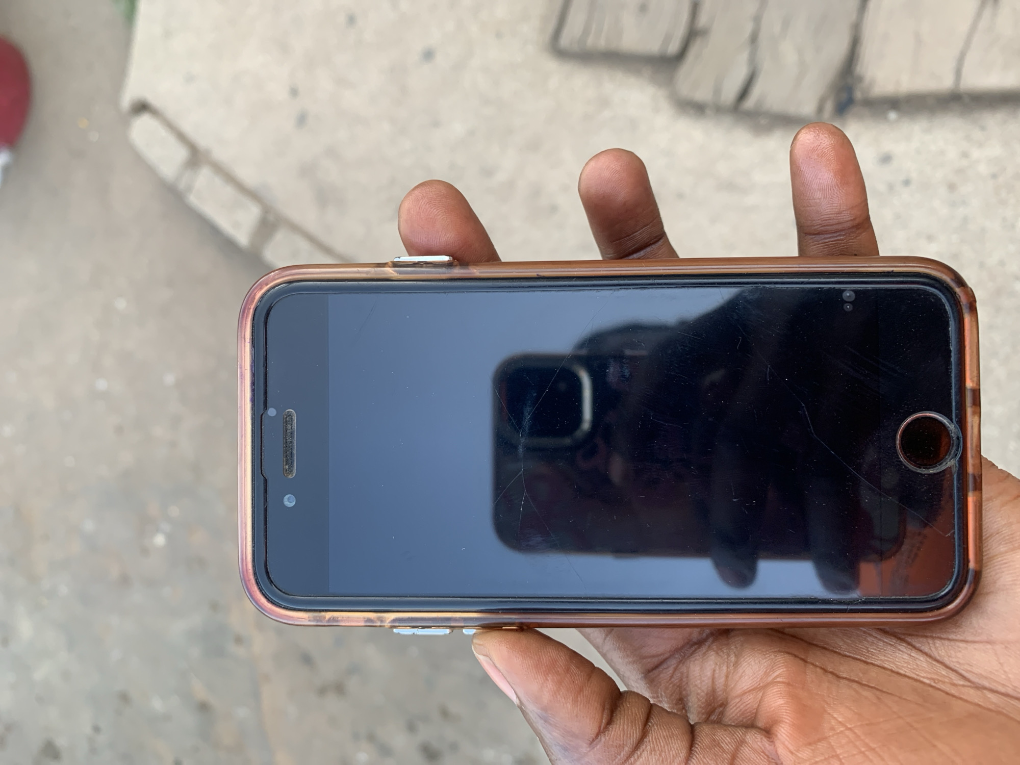 A dark-skinned hand holds a cracked iPhone in a brown case. The phone's screen is reflecting the surroundings. The location of New Stadium, Nigeria, cannot be determined from the visual information provided.