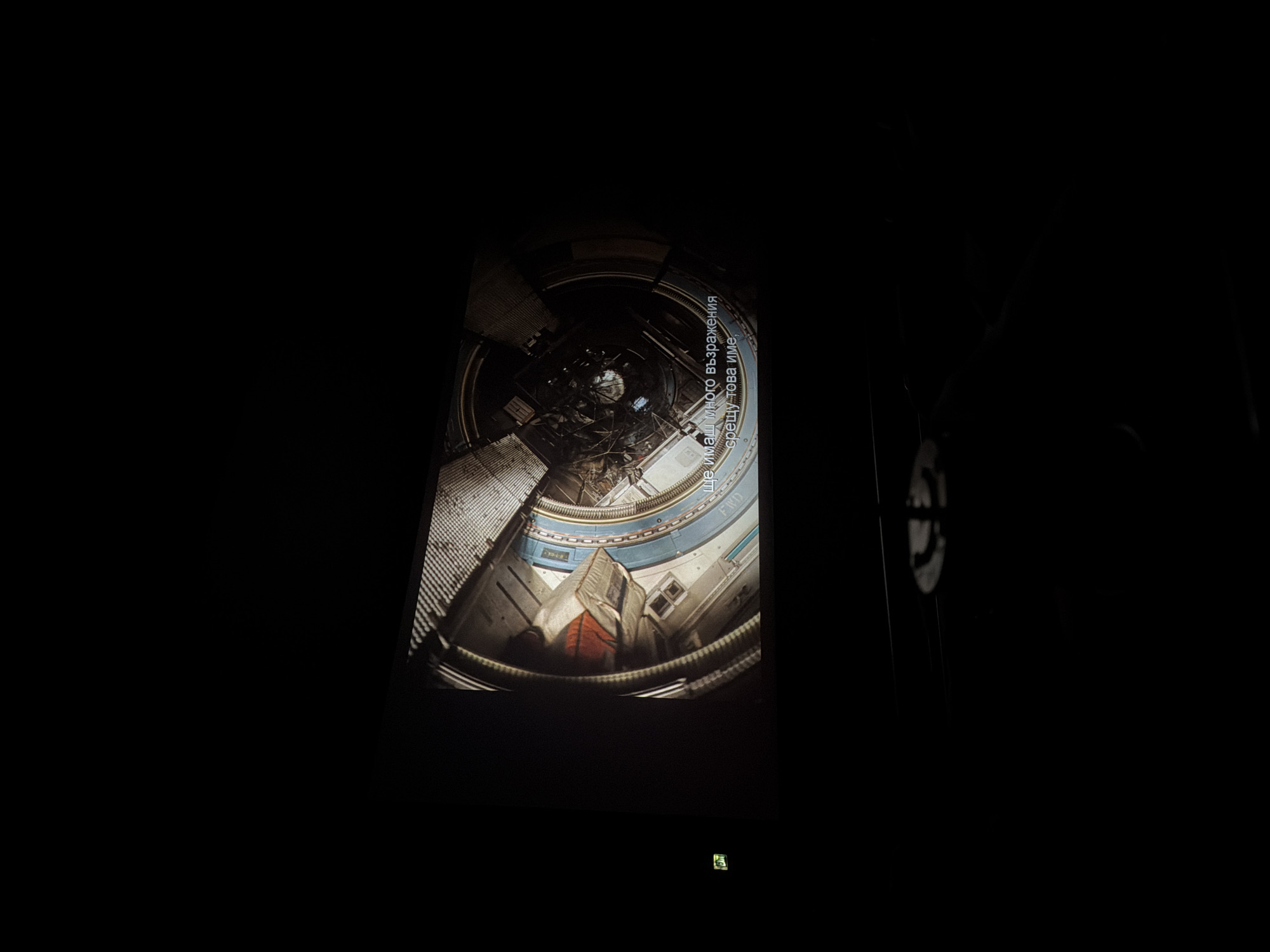 The image shows a screen displaying a scene inside what appears to be a space station or spacecraft interior, with a view looking upwards. Text in Bulgarian is visible on the screen. The image is taken in a dark environment with an emergency exit sign visible at the bottom of the frame. The exact location in Sofia, Bulgaria, cannot be determined from the image.