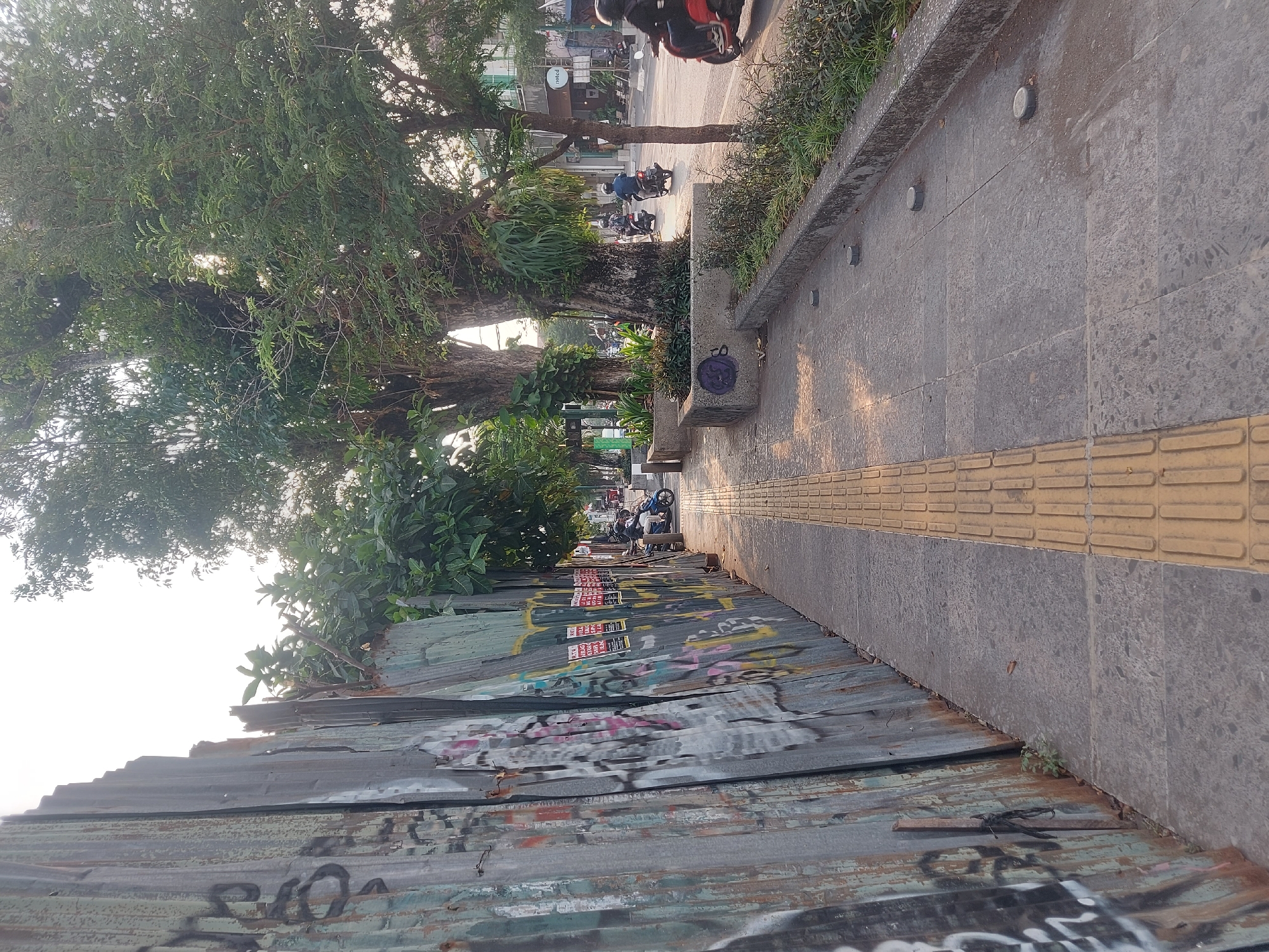 A paved sidewalk with tactile paving for the visually impaired runs alongside a weathered corrugated metal fence covered in graffiti. The fence is on the left side of the image, and the sidewalk is on the right. Trees with lush green foliage line the sidewalk, casting dappled shadows. In the background, a street is visible with several motorbikes driving by. The scene appears to be in an urban area, likely in Yogyakarta, Indonesia, given the presence of motorbikes and the architectural style of the buildings in the distance. The lighting suggests it is either early morning or late afternoon.