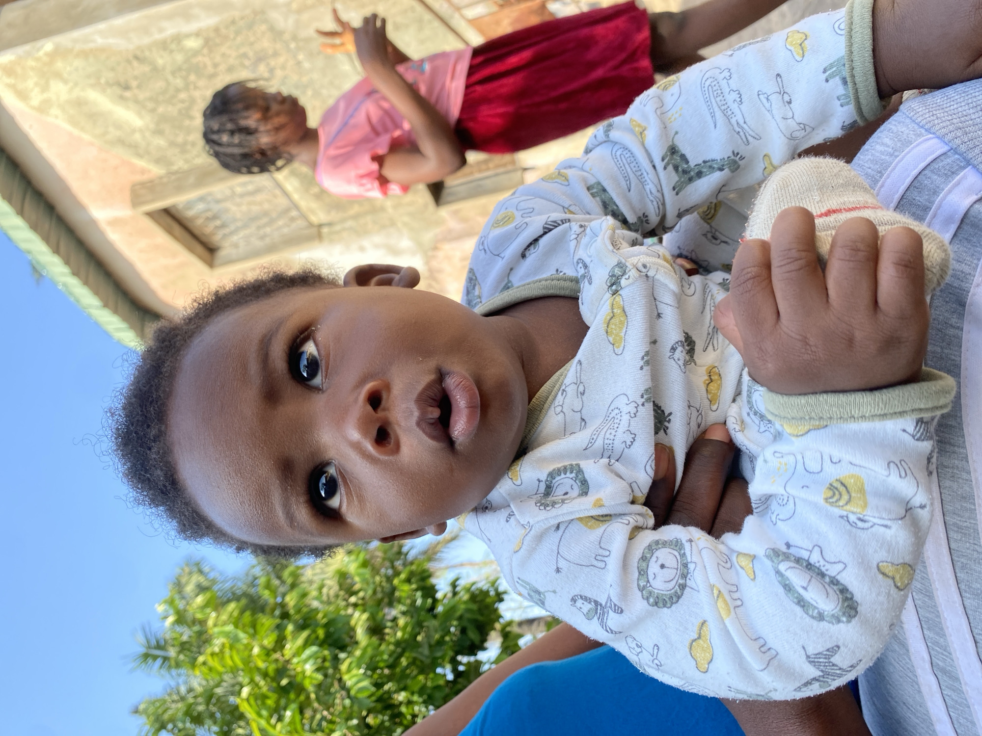 In Sabon Tasha, Nigeria, a close-up, low-angle shot features a dark-skinned baby held by an unseen person. The baby, wearing a white onesie with cartoon animal prints, looks upwards with wide eyes and an open mouth. To the right, in the background, a young girl with braided hair and wearing a pink shirt and red skirt is blurred. The environment appears to be outdoors with a building and greenery visible.