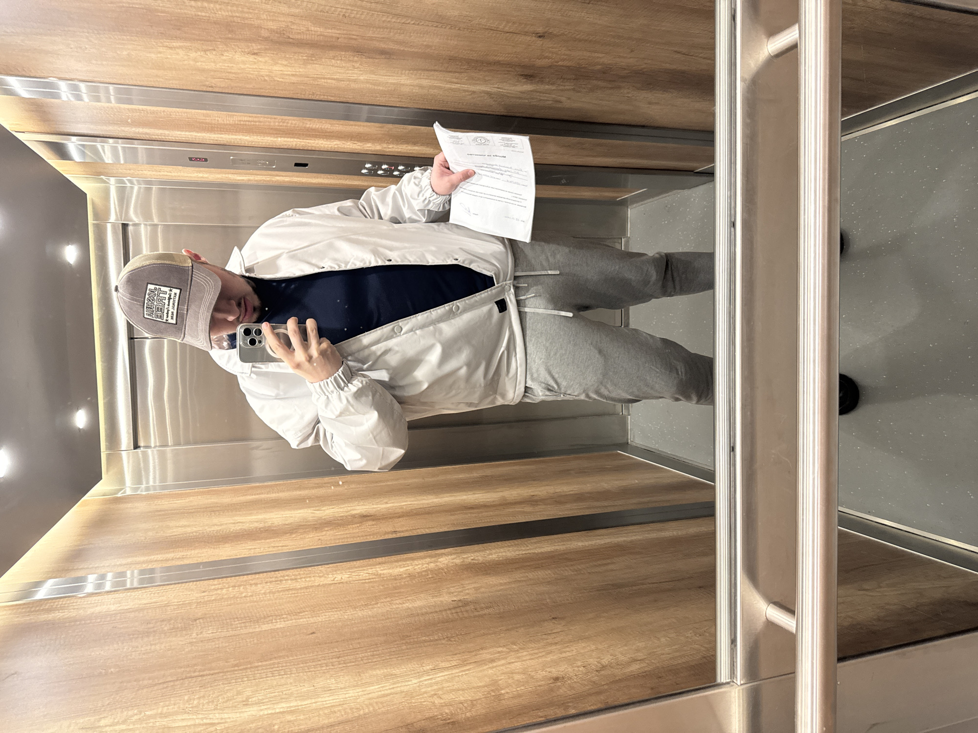 The image captures a person taking a mirror selfie inside a modern elevator. The individual, dressed in a light grey or white windbreaker jacket over a dark blue t-shirt, grey sweatpants, and a grey and beige trucker-style baseball cap, stands facing their reflection. They hold a grey smartphone in their left hand, aimed at the mirror for the selfie, and a white document in their right hand.
The elevator's interior features a blend of materials. One side wall and part of the back wall are clad in light brown wood-effect panels. The opposite wall, where the person is reflected, and the door frame are made of polished stainless steel, creating a mirror-like surface. A sturdy, silver-colored handrail runs vertically and horizontally along the mirrored wall. The floor is a dark grey, textured material. Recessed lighting elements are visible on the ceiling, casting bright reflections on the metallic surfaces. Elevator control buttons are visible on a panel on the wooden wall, though specific labels are indistinguishable.
Notable details include the mirrored text on the person's cap, which appears to be a brand logo or slogan, possibly reading "ZEMAT" and "WELLDEY. WORK" with "102HM" below it. The document held by the person also shows mirrored text; a larger heading at the top is partially visible, potentially reading "MoniuM ex SonobM", followed by several lines of smaller, illegible text. The person's expression is neutral, focused on the phone. The setting suggests an indoor, contemporary environment, likely a residential or commercial building.
