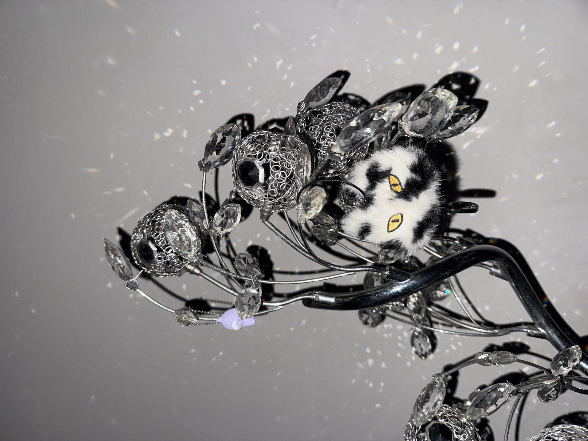 The image displays a close-up of an ornate decorative light fixture, likely a chandelier, set against a plain, light-grey background. The fixture is intricate, featuring numerous clear, multi-faceted crystals of various shapes and sizes that reflect and refract light, creating a sparkling effect. Interspersed among the crystals are several spherical elements crafted from intricate, filigree-like metalwork, possibly silver-colored, with some exhibiting dark, reflective centers.
A distinctive black and white furry pom-pom or charm, adorned with two prominent yellow, almond-shaped eyes with black pupils, resembling animal eyes, is attached to the fixture. Below this, a small, light purple charm, possibly made of plastic or ceramic and shaped like a simplified figure, hangs delicately. The main structure of the fixture comprises slender silver-colored wires and a thicker, dark, curved metallic branch from which these decorative elements extend.
The most striking visual element is the myriad of bright, iridescent specks of light scattered across the light-grey background, which are reflections and refractions from the crystals. These sparkling highlights suggest a brightly lit environment, likely indoors during the day. No human activity or people are visible in the static scene. There is no discernible text. The decorative piece is situated in a residential setting at บ้านกาญจน์กนก 23, Thailand, contributing an enchanting and whimsical atmosphere.
