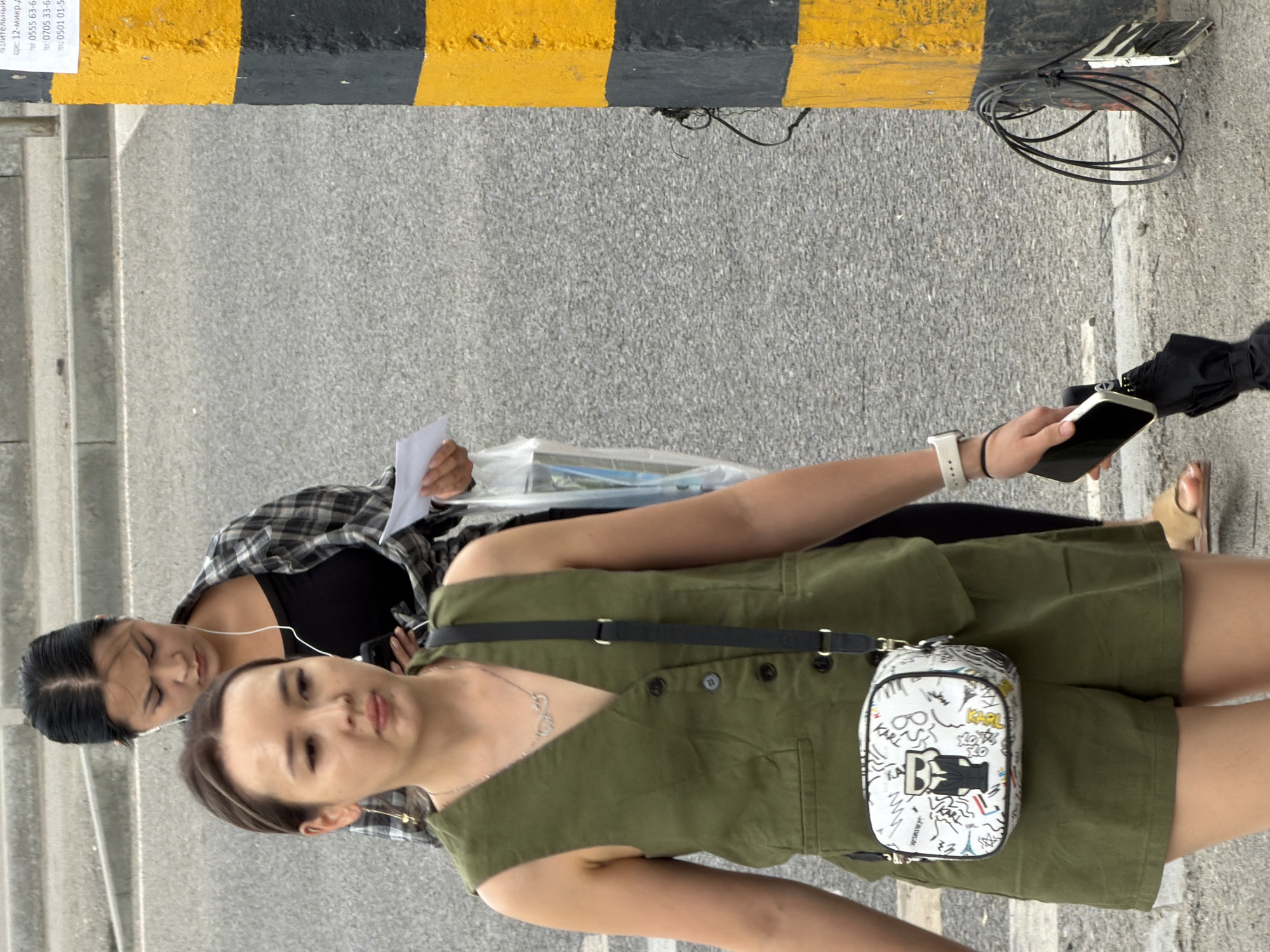Two women are walking on a paved surface, possibly a street or sidewalk. The woman in the foreground, facing left, is wearing a green sleeveless vest over a green outfit and a white crossbody bag decorated with cartoonish figures and the name "KARL". She has earrings, a necklace, and a white watch on her left wrist. She is holding a black smartphone in her right hand and an open black umbrella in her left. The woman in the background, facing forward, is wearing a black and gray plaid shirt over a black top and has earphones in. She is holding a white piece of paper in her right hand. A black and yellow striped pole is visible on the right side of the image. The ground is asphalt with some concrete curbs.